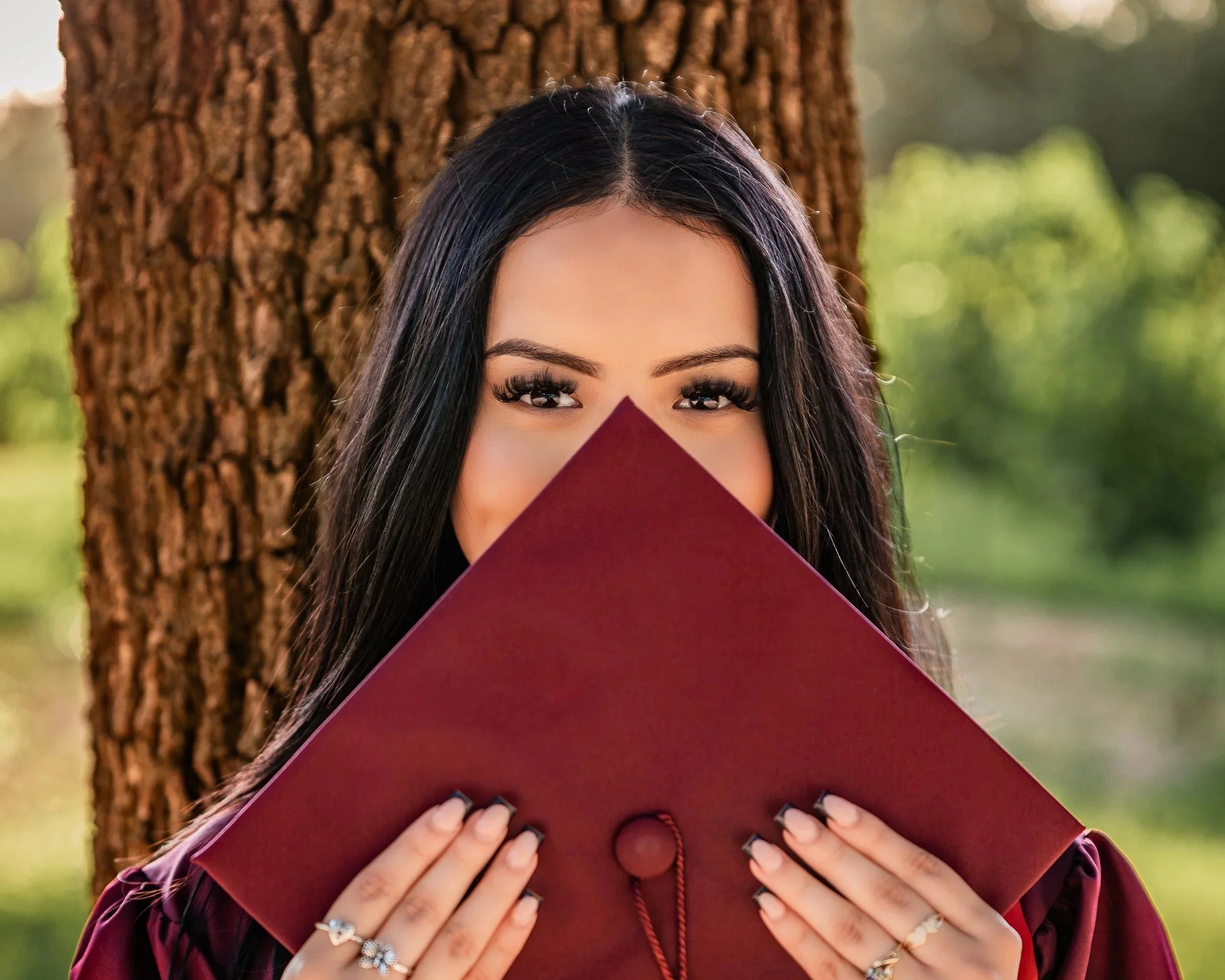 A young woman with long dark hair holding a maroon diploma cover in front of her face, standing outdoors with a tree in the background.