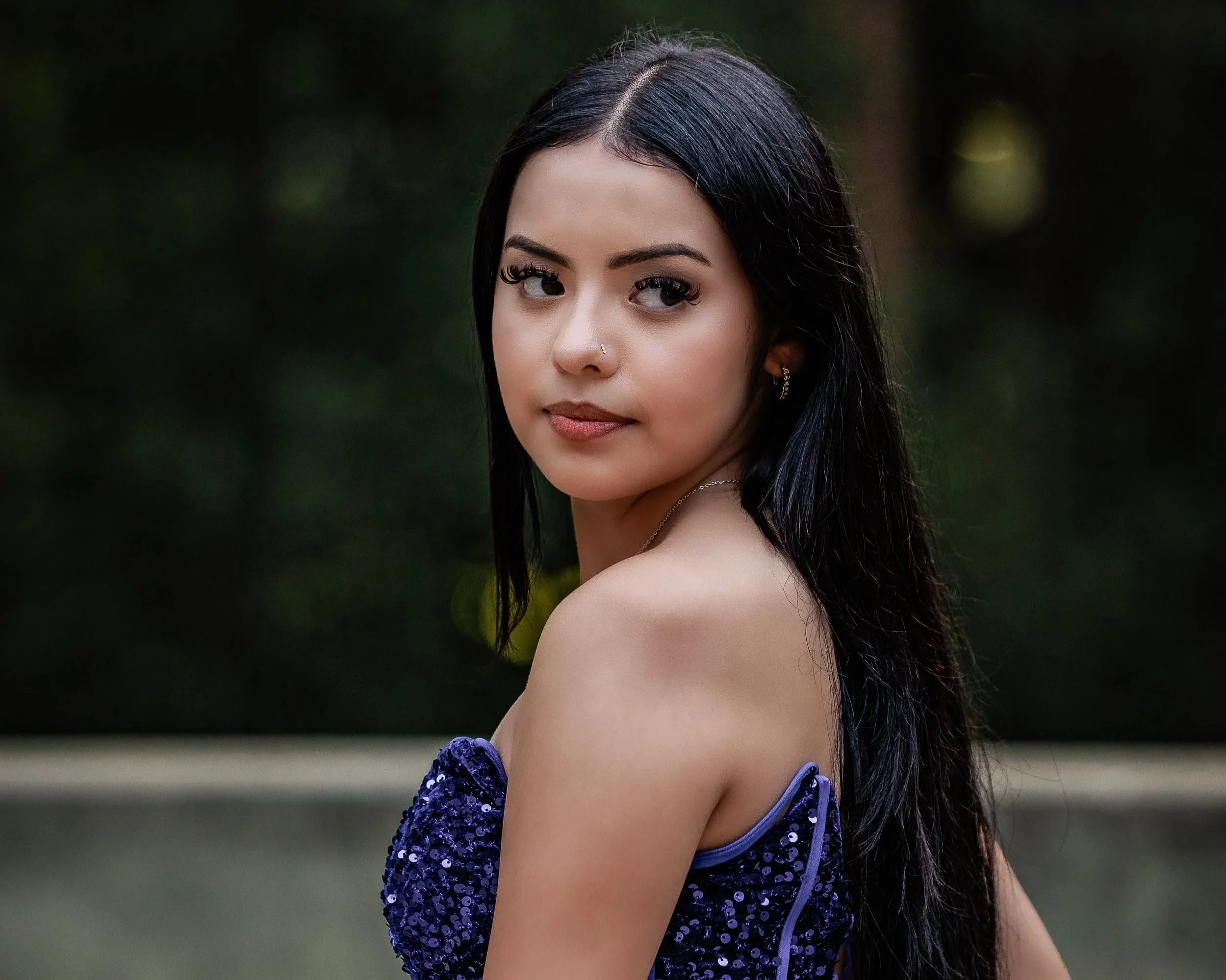 A young woman with long black hair and makeup, wearing a purple sequin dress, looking over her shoulder outdoors with a blurred green background.