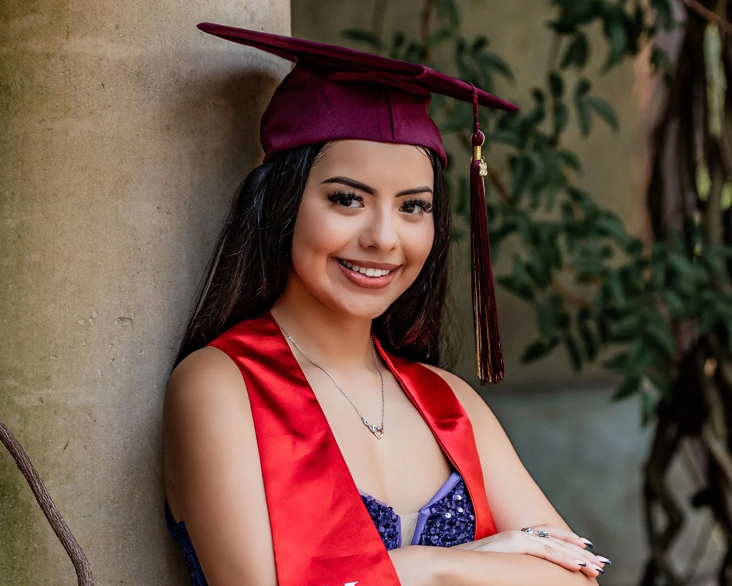 Young woman in a maroon graduation cap and red stole, smiling, with dark hair and makeup, standing outdoors against a wall with green foliage in the background.