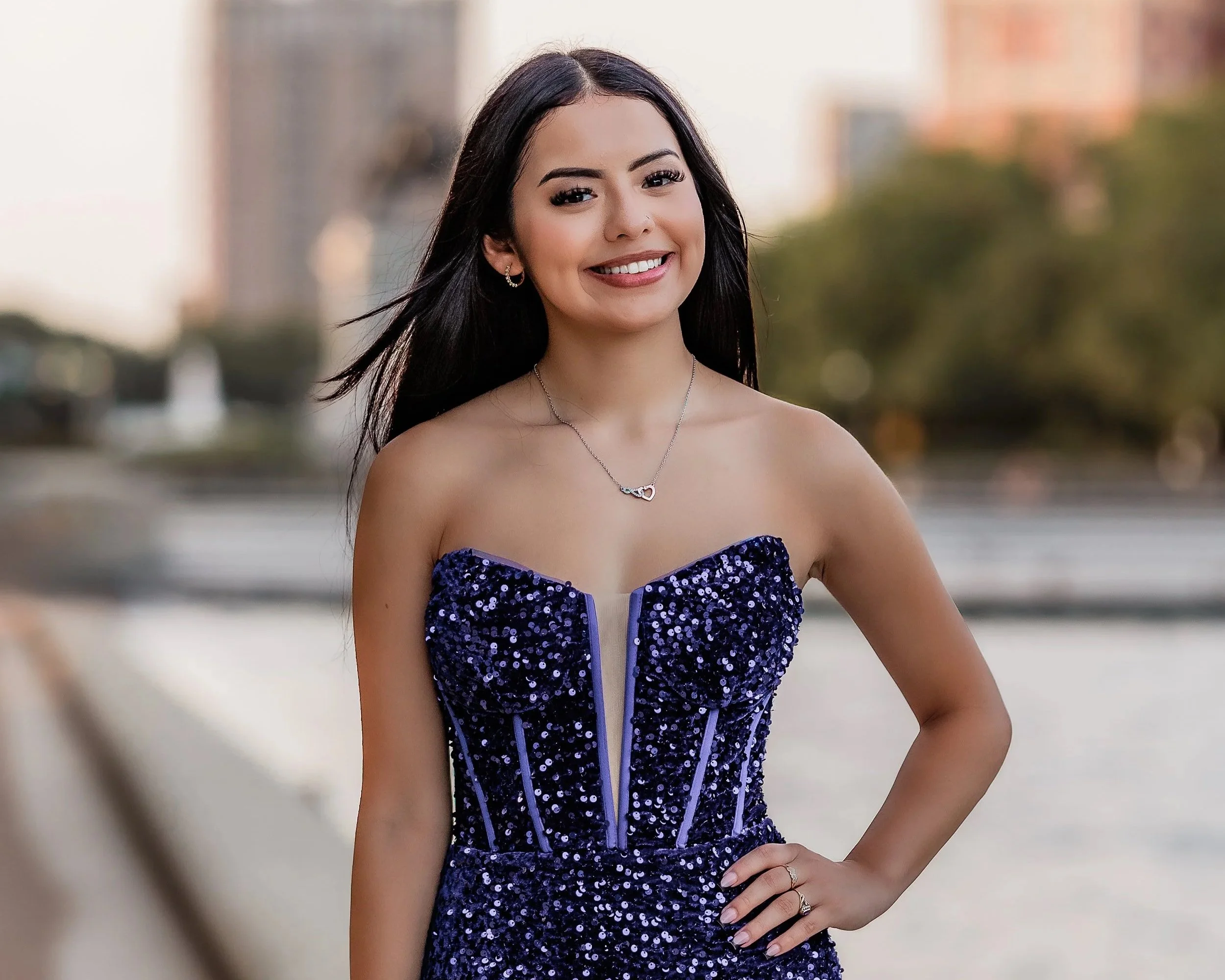 A young woman with long black hair smiling outdoors, wearing a strapless, dark blue, sequined dress with a necklace and ring.