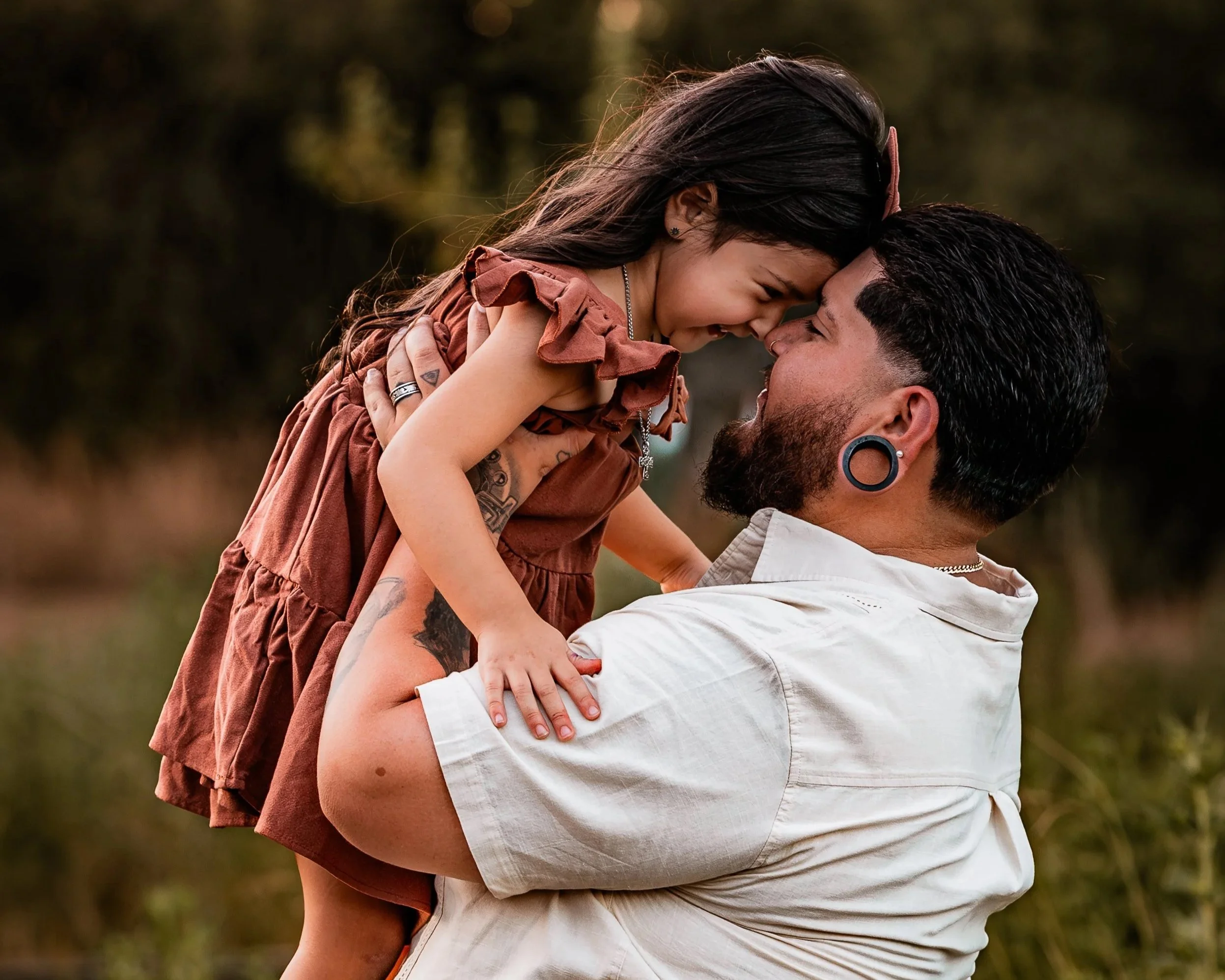 A man with a beard and large gauge earrings lifting a young girl, both smiling and touching foreheads outdoors in nature.