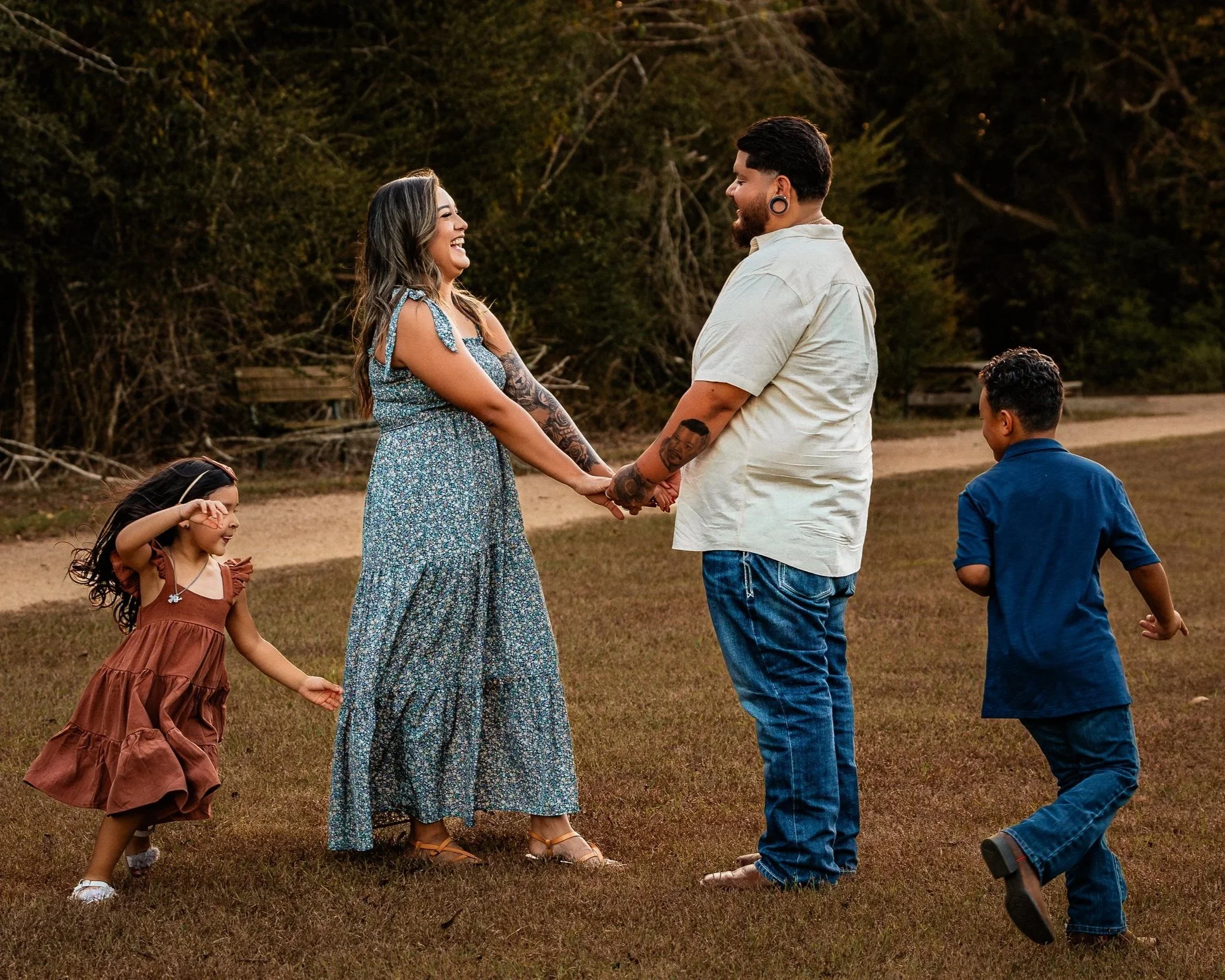 A couple holding hands and smiling at each other in a grassy outdoor area with two children playing nearby.