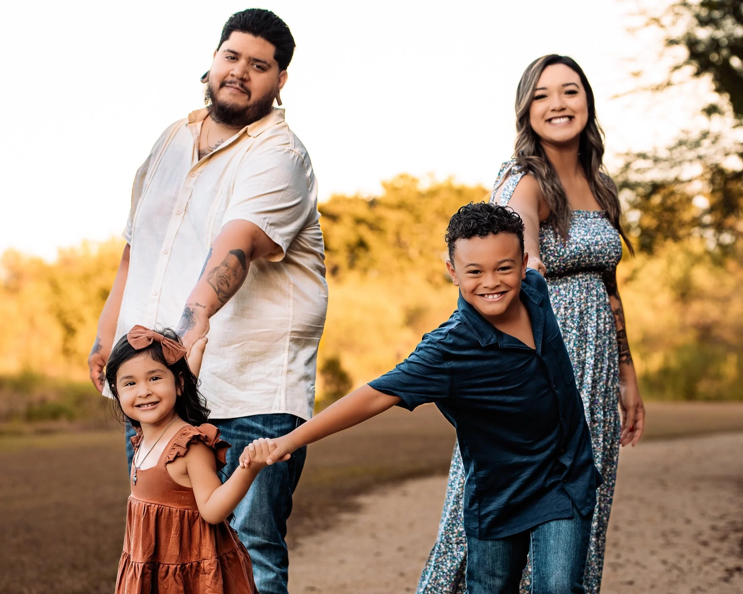 A diverse family posing outdoors during golden hour. The father is holding hands with a young girl, and a young boy is playfully leaning back, smiling. The mother is smiling in the background. They are in a natural setting with trees and an open sky.