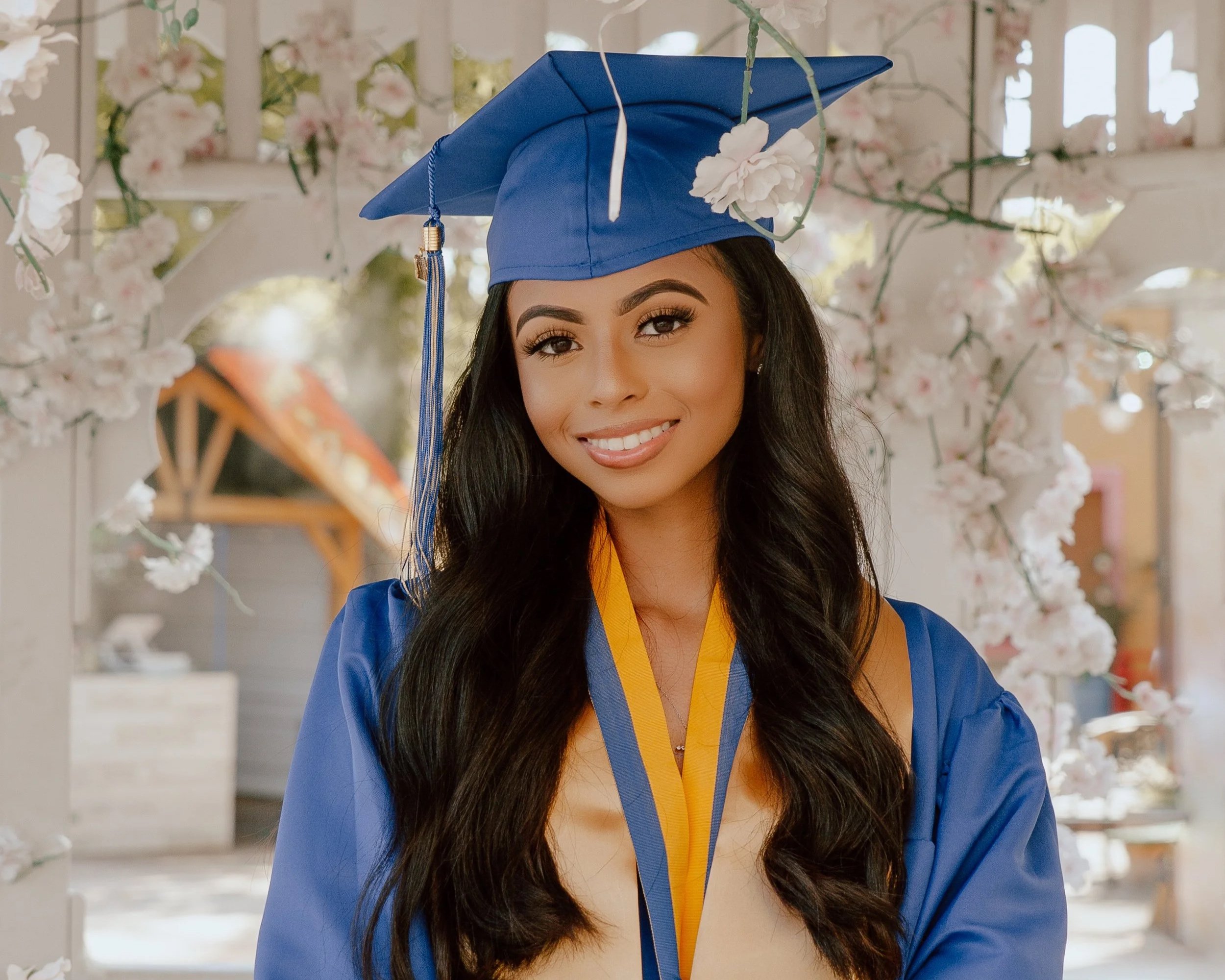 A young woman with long dark hair and makeup wearing a blue graduation cap and gown with a yellow sash, smiling in front of a floral backdrop.
