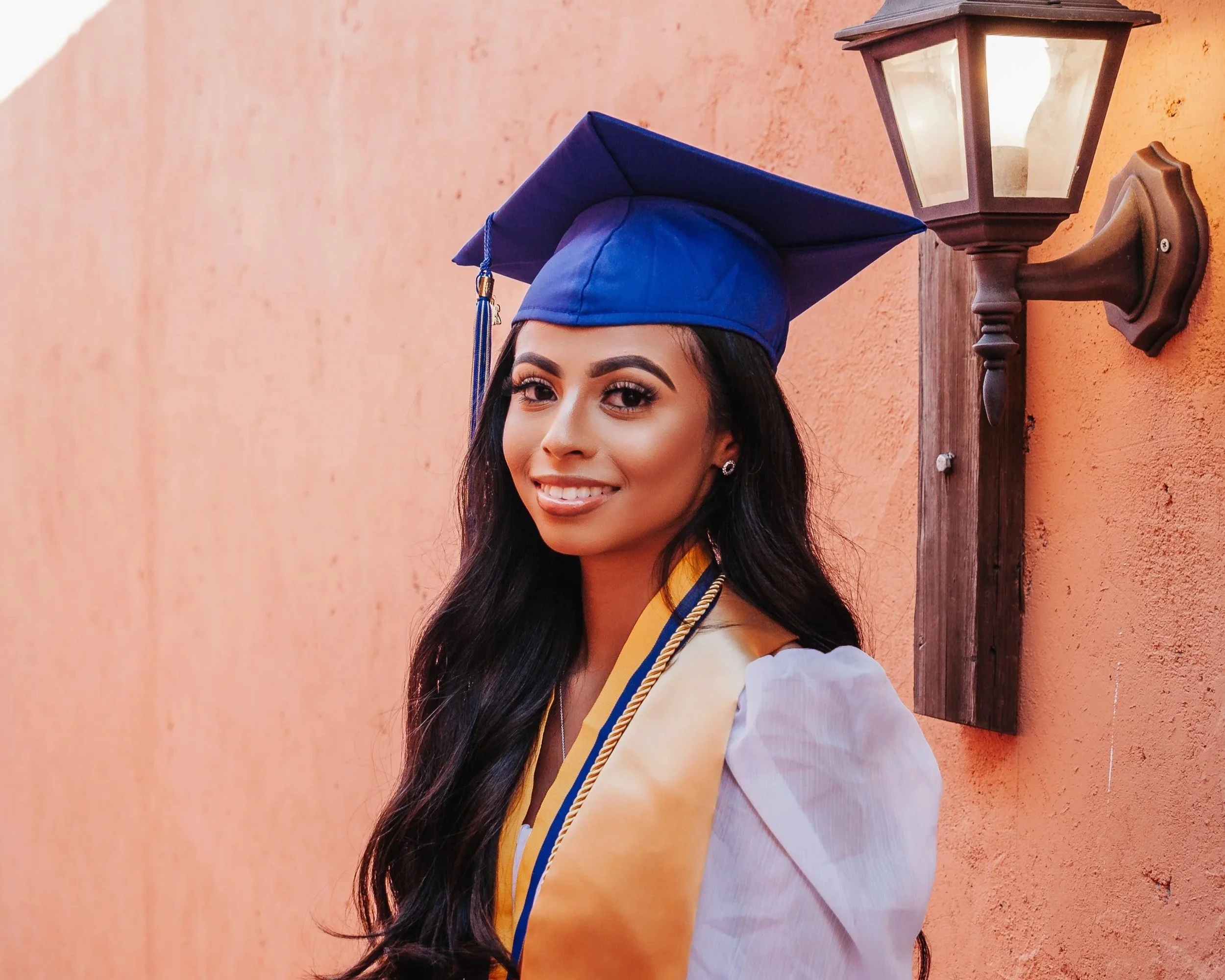 A young woman in graduation attire, with a blue cap and a yellow stole, smiling while standing in front of a peach-colored wall with a wall-mounted lantern.