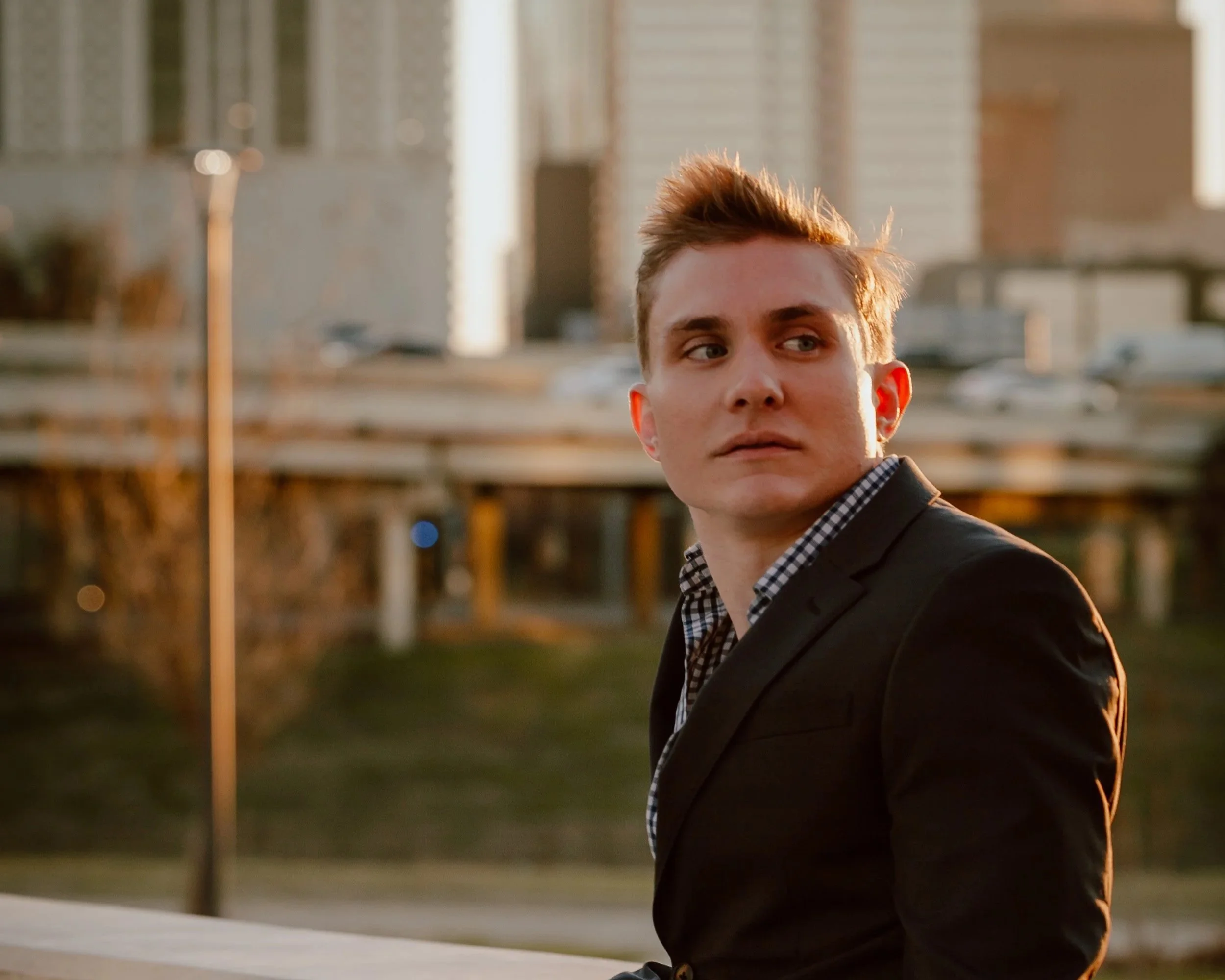 A young man with light brown hair wearing a black jacket and checkered shirt, looking off to the side, outdoors at sunset with a blurred cityscape in the background.