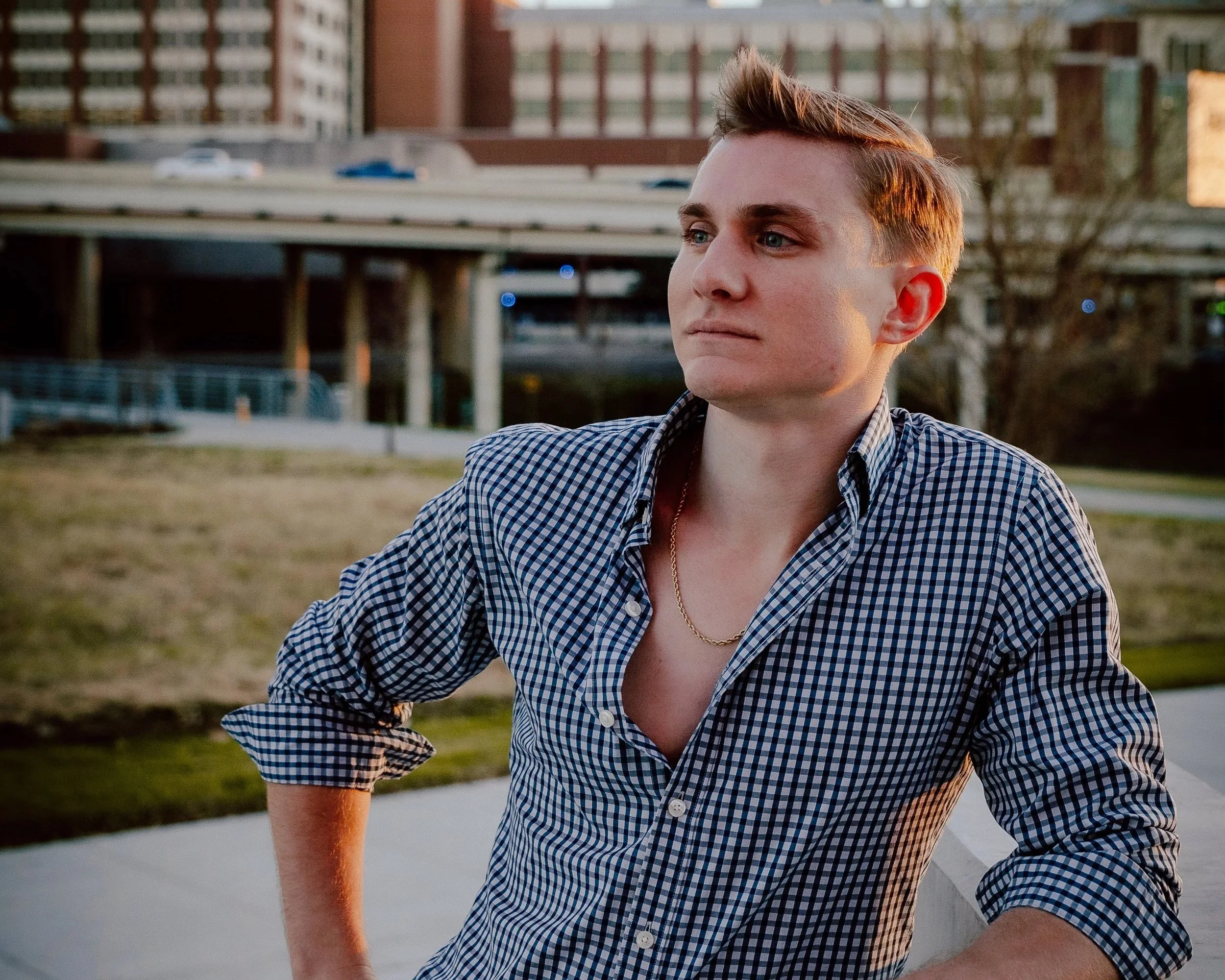Young man with short hair and a checkered shirt standing outdoors in an urban park during sunset.