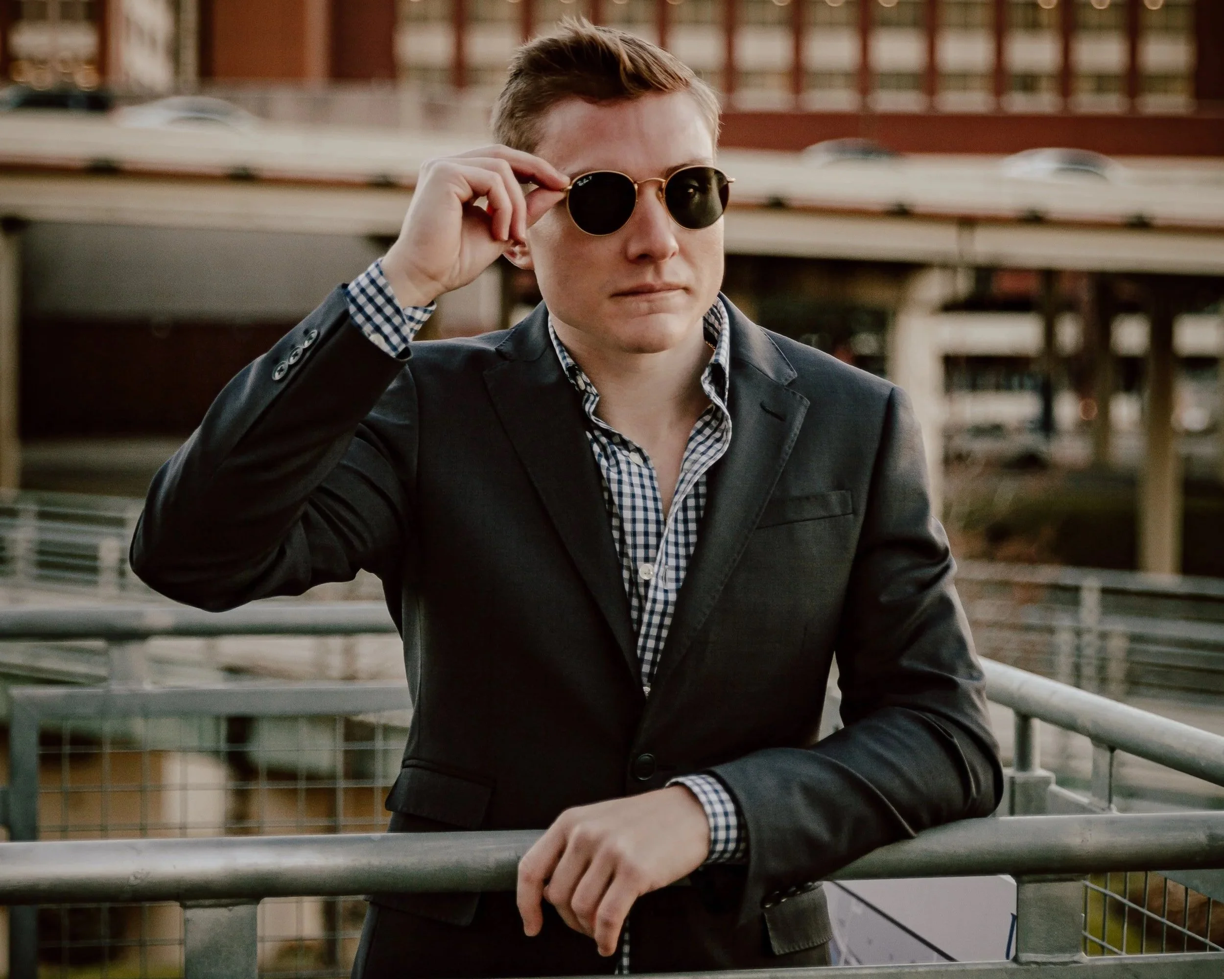 A man in a black suit and checkered shirt wearing sunglasses, standing on a balcony with a cityscape in the background.