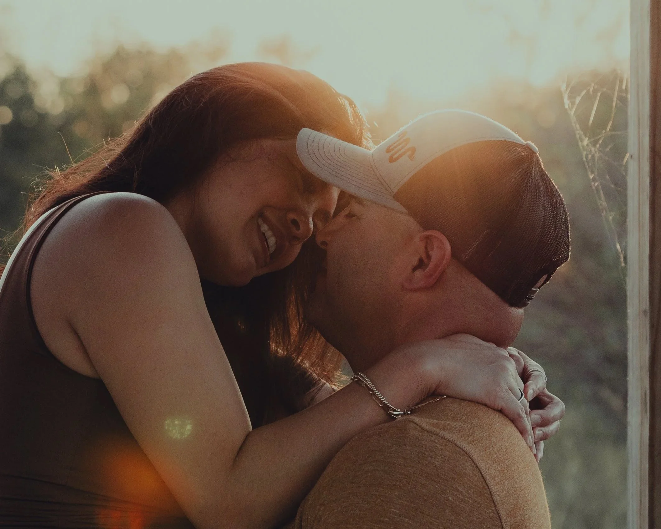 A couple sharing a close, intimate moment, touching foreheads and smiling at each other, with sunlight in the background.