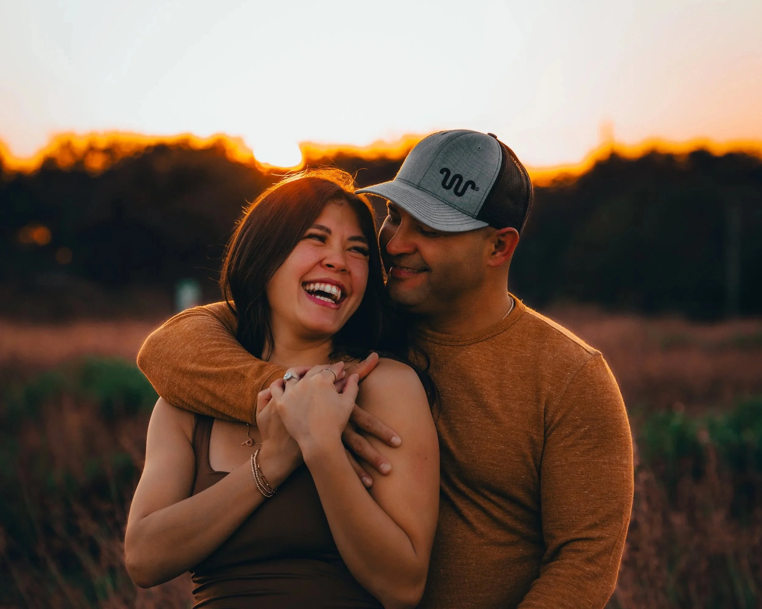 A couple smiling and hugging outdoors at sunset, with the warm glow of the sun behind them.