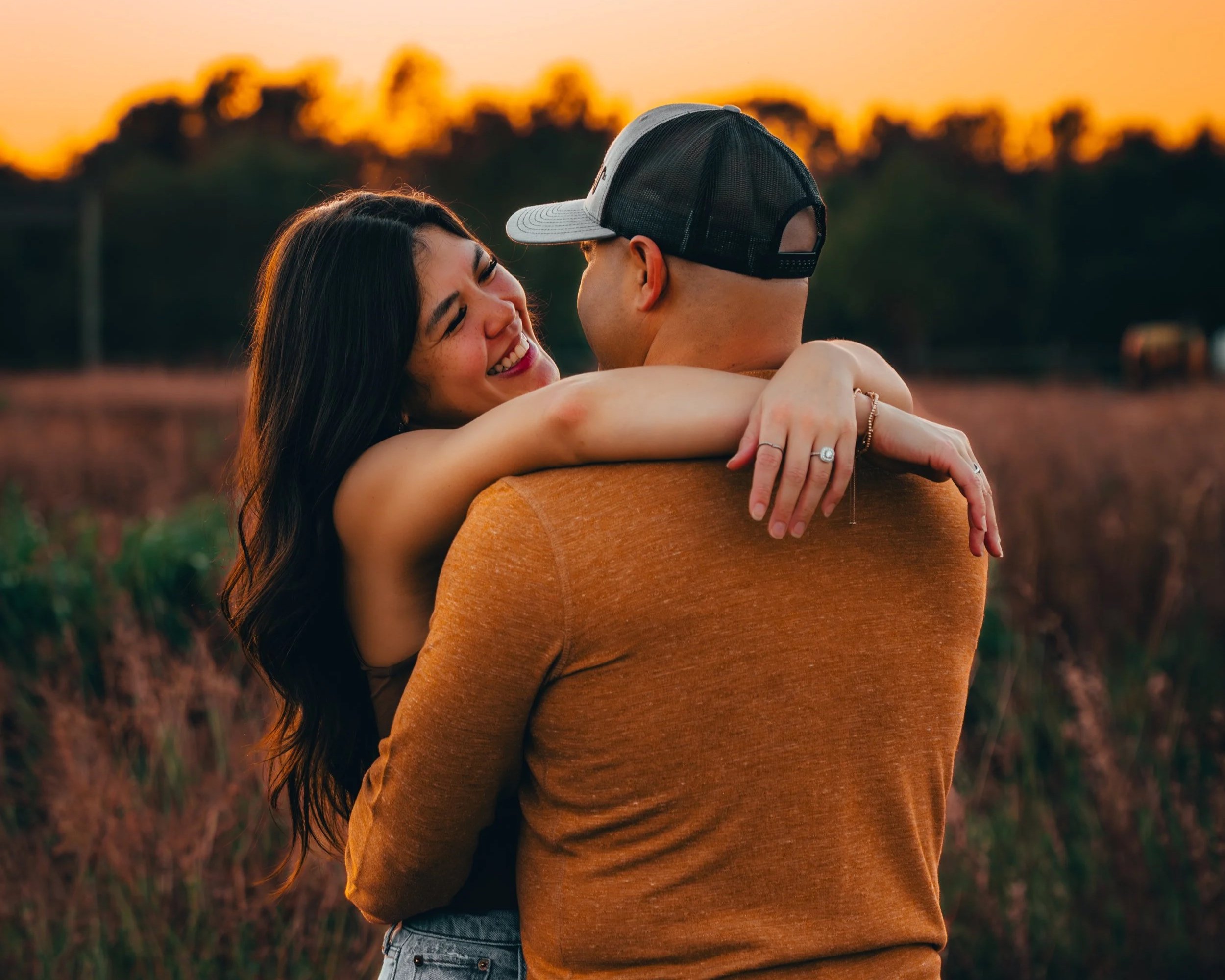 A smiling woman and man embrace in a field at sunset, with the woman looking joyful and the man wearing a baseball cap.