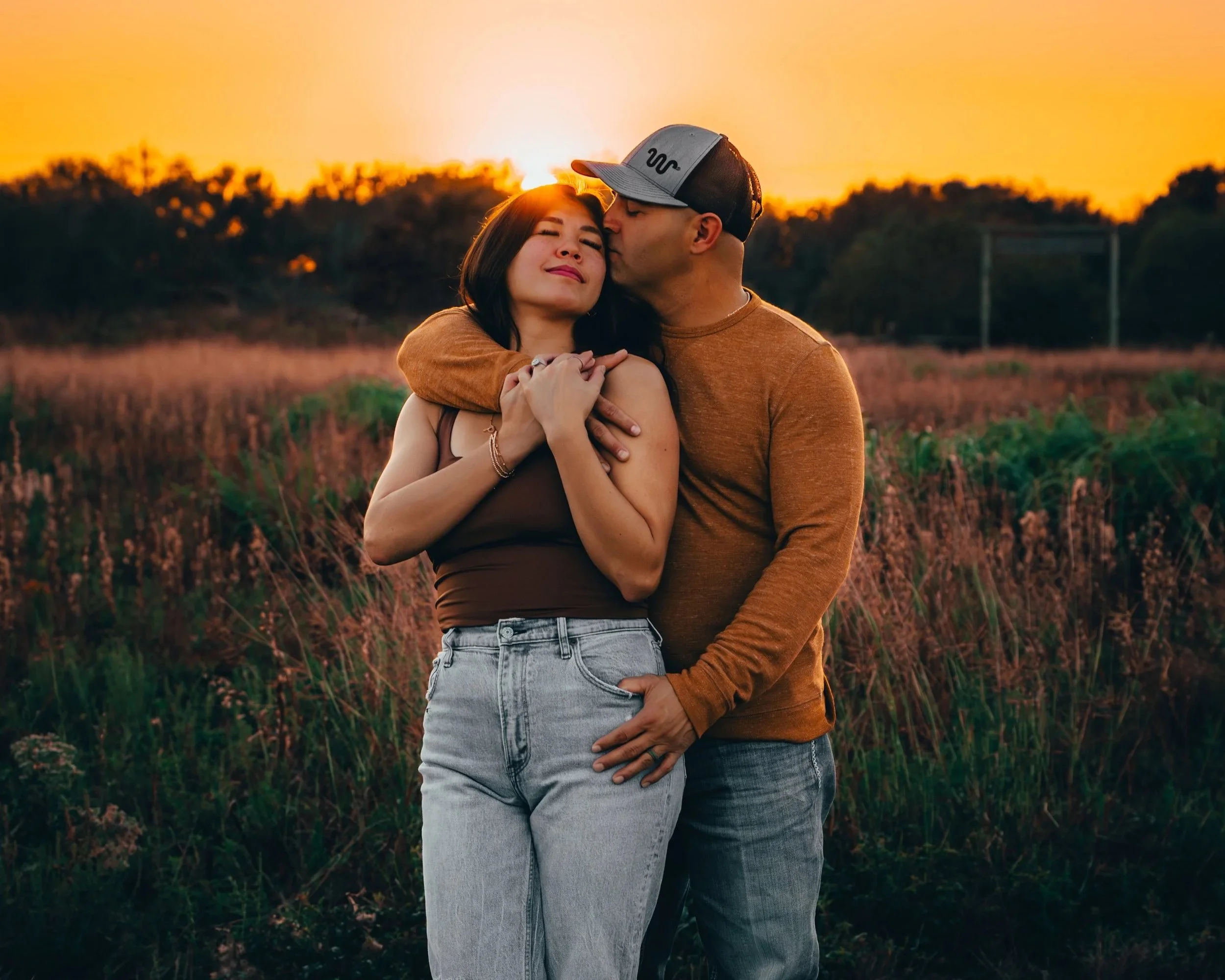 A couple embracing outdoors at sunset, the man kissing the woman on the cheek. She has her eyes closed and their arms are around each other, standing in a field with trees in the background.