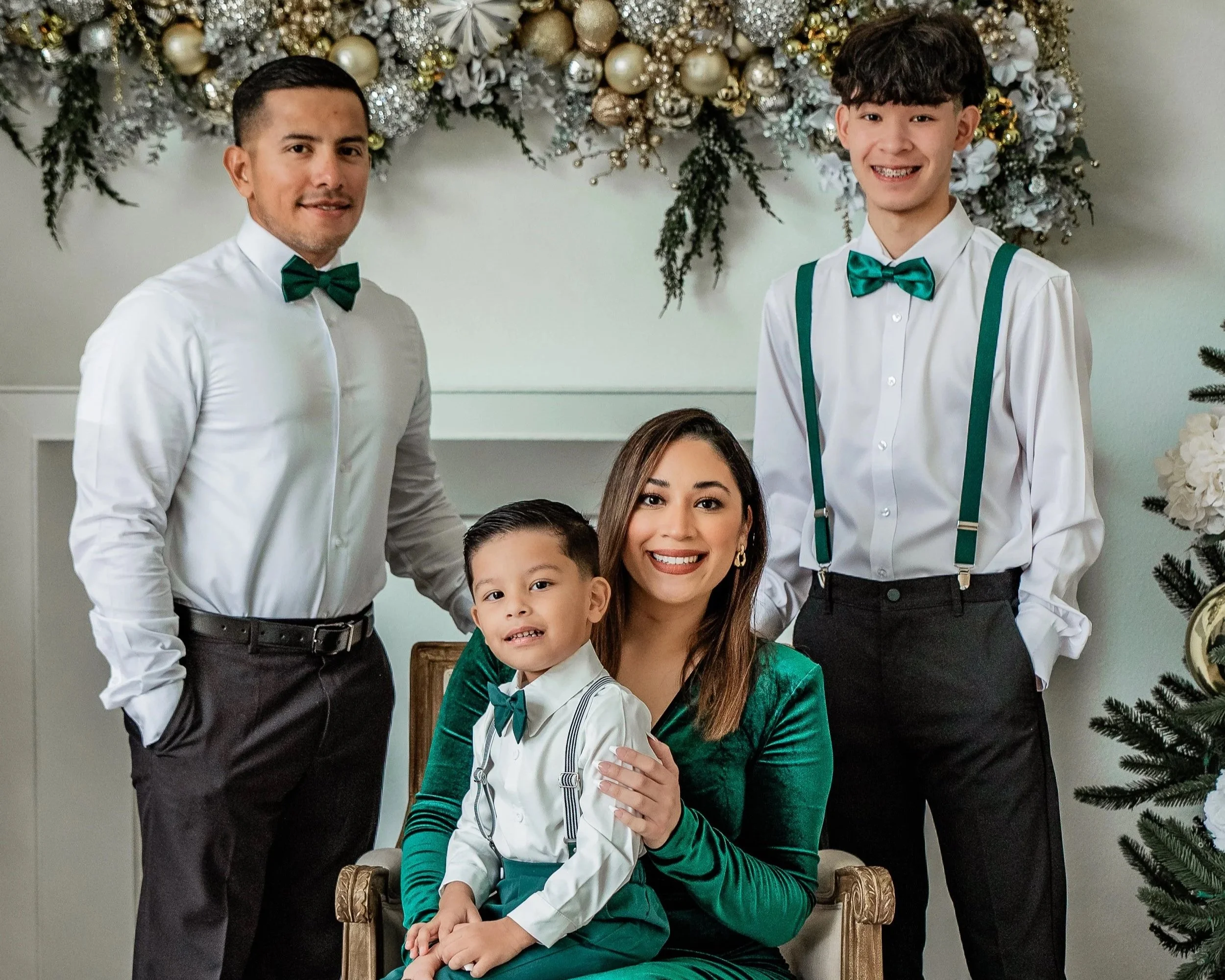 A family of five dressed in festive holiday attire, posing indoors with Christmas decorations including a decorated tree and a garland, smiling at the camera.