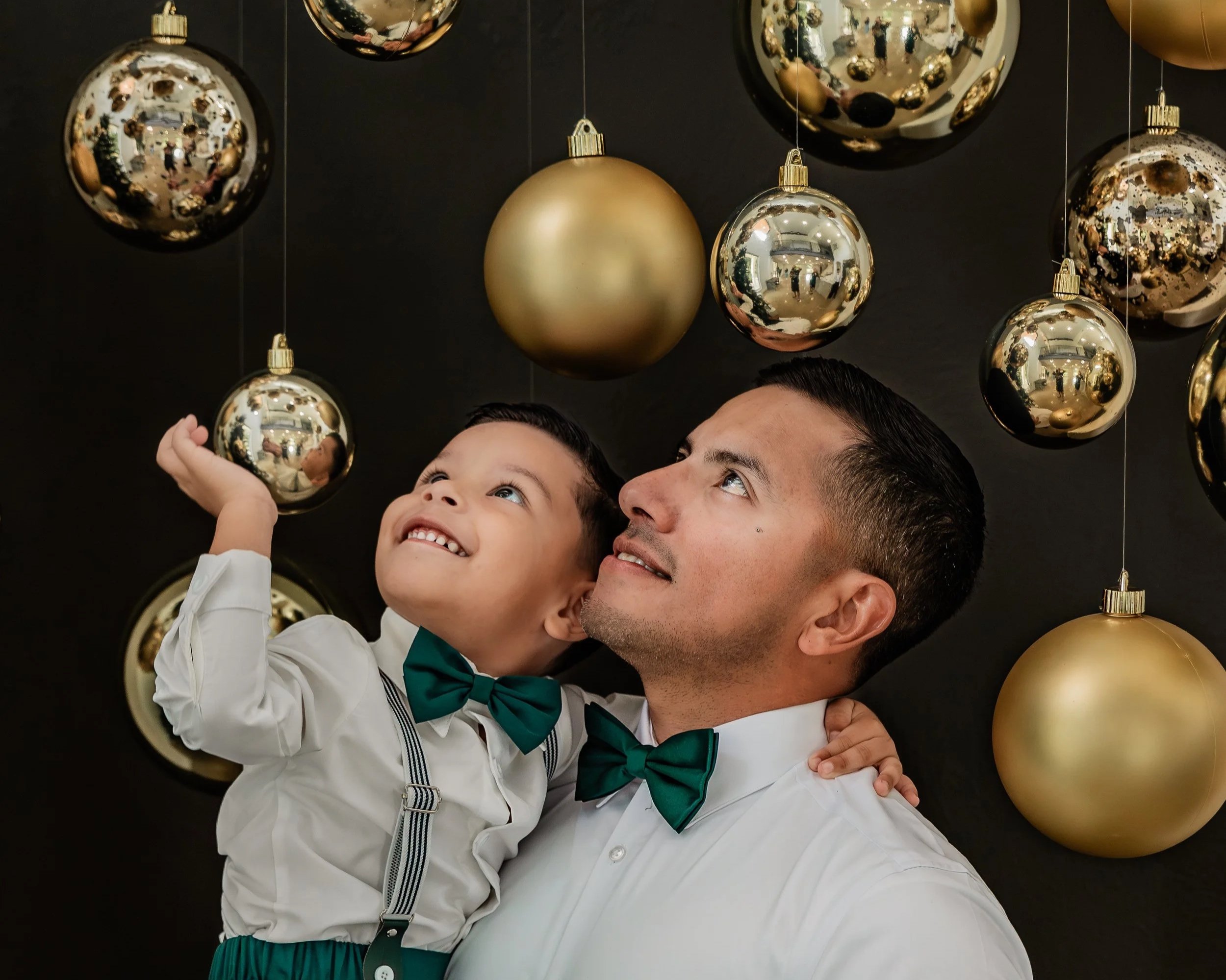 A man and a young boy smiling and looking up at hanging gold and silver Christmas ornaments, both dressed in white shirts and green bow ties, with the boy also wearing suspenders.