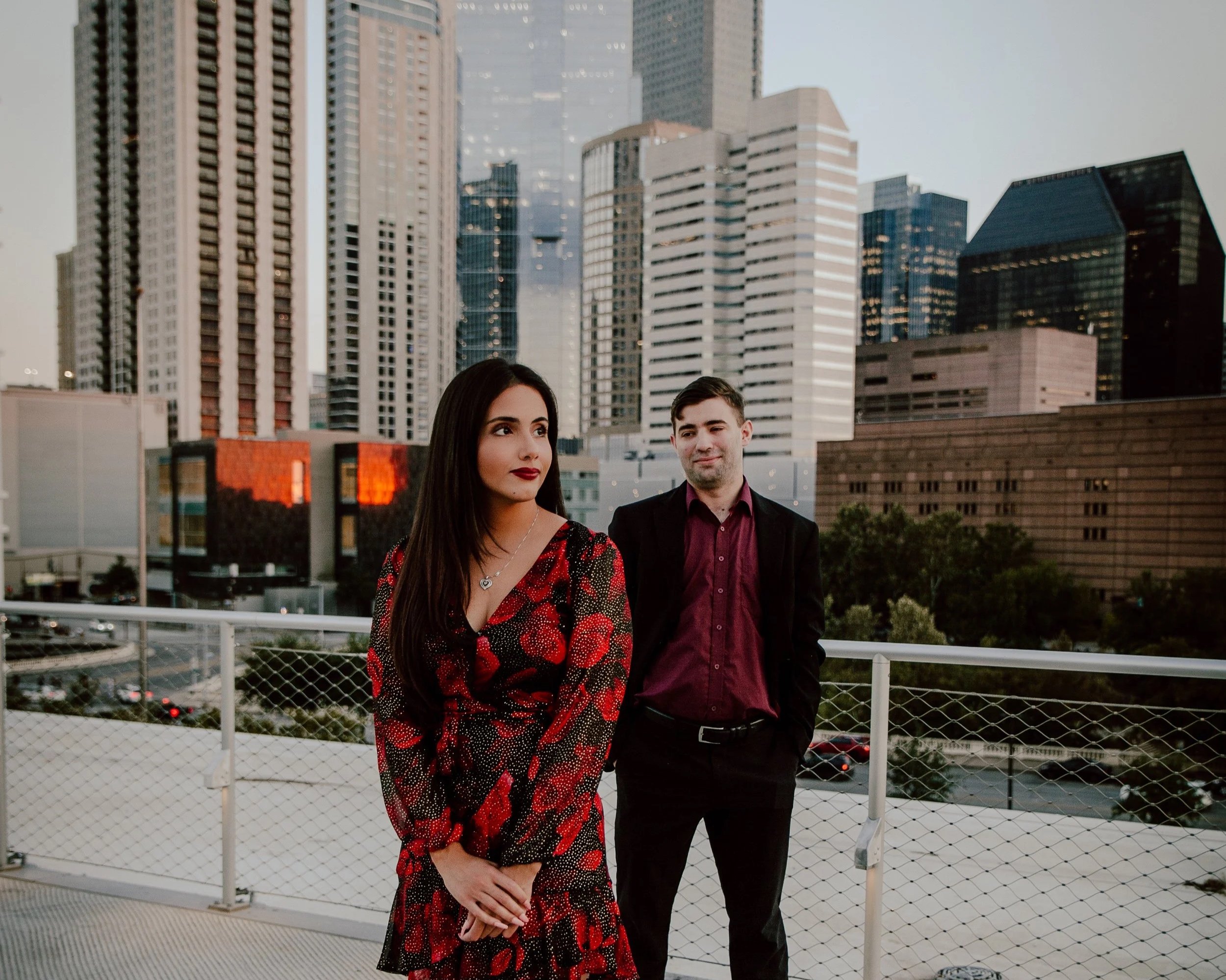 A woman and a man stand on a rooftop with a city skyline in the background during sunset. The woman wears a red and black floral dress, and the man wears a dark suit with a maroon shirt.