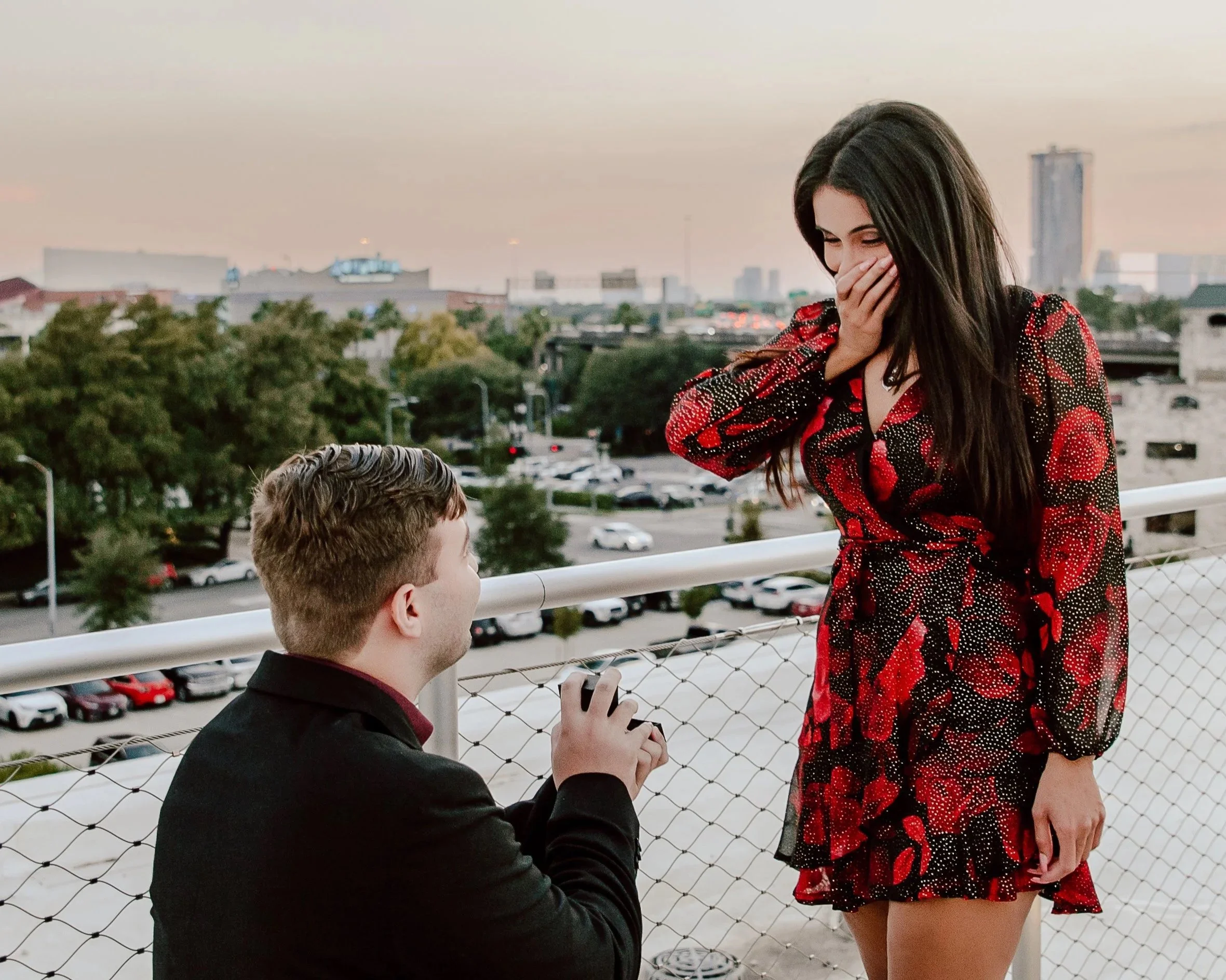A man proposes to a woman on a rooftop overlooking a cityscape at sunset. The woman is surprised and covering her mouth with her hand, wearing a black and red floral dress. The man is kneeling, holding an open ring box.