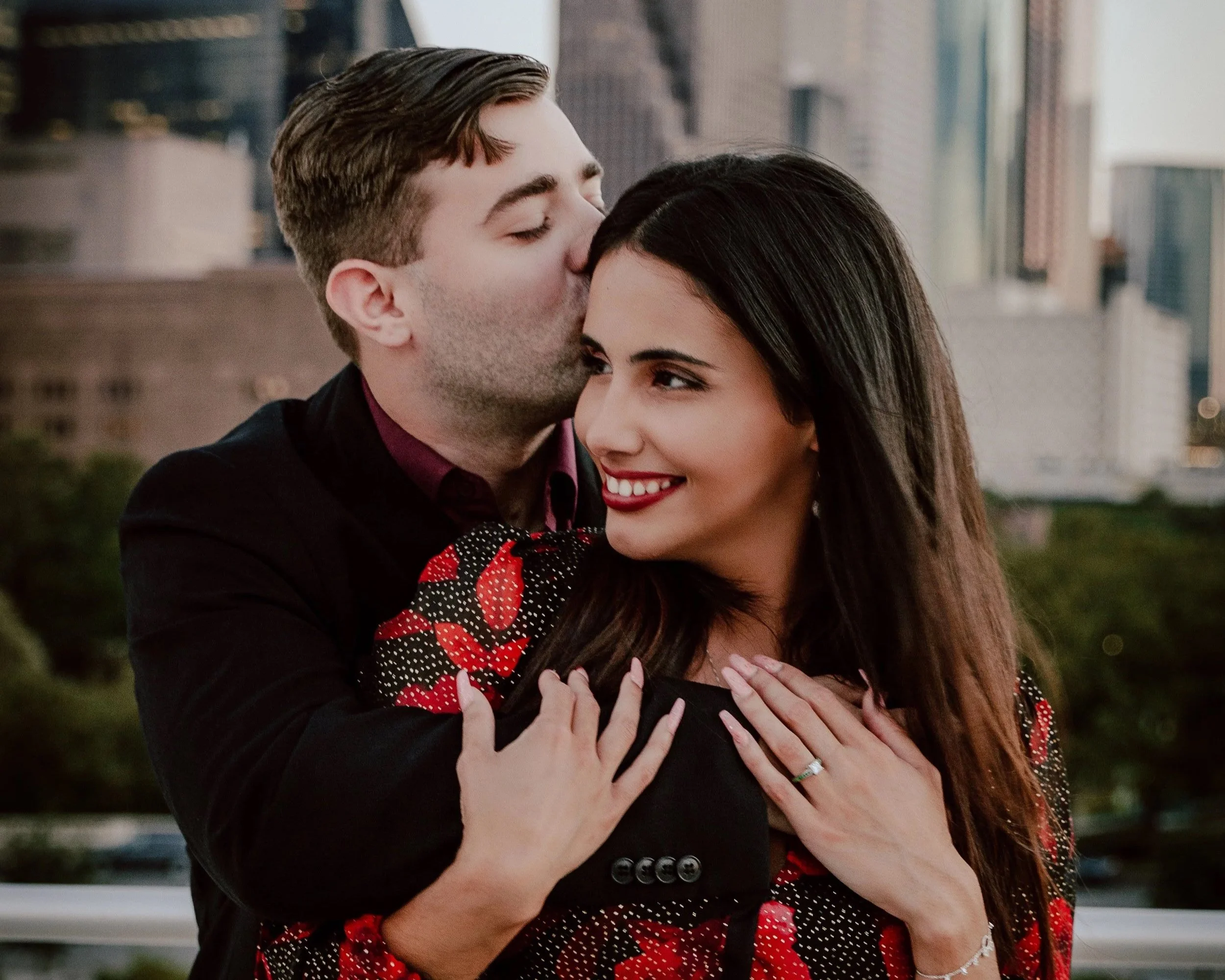 A man kisses a woman's forehead as she smiles warmly, with a city skyline in the background.
