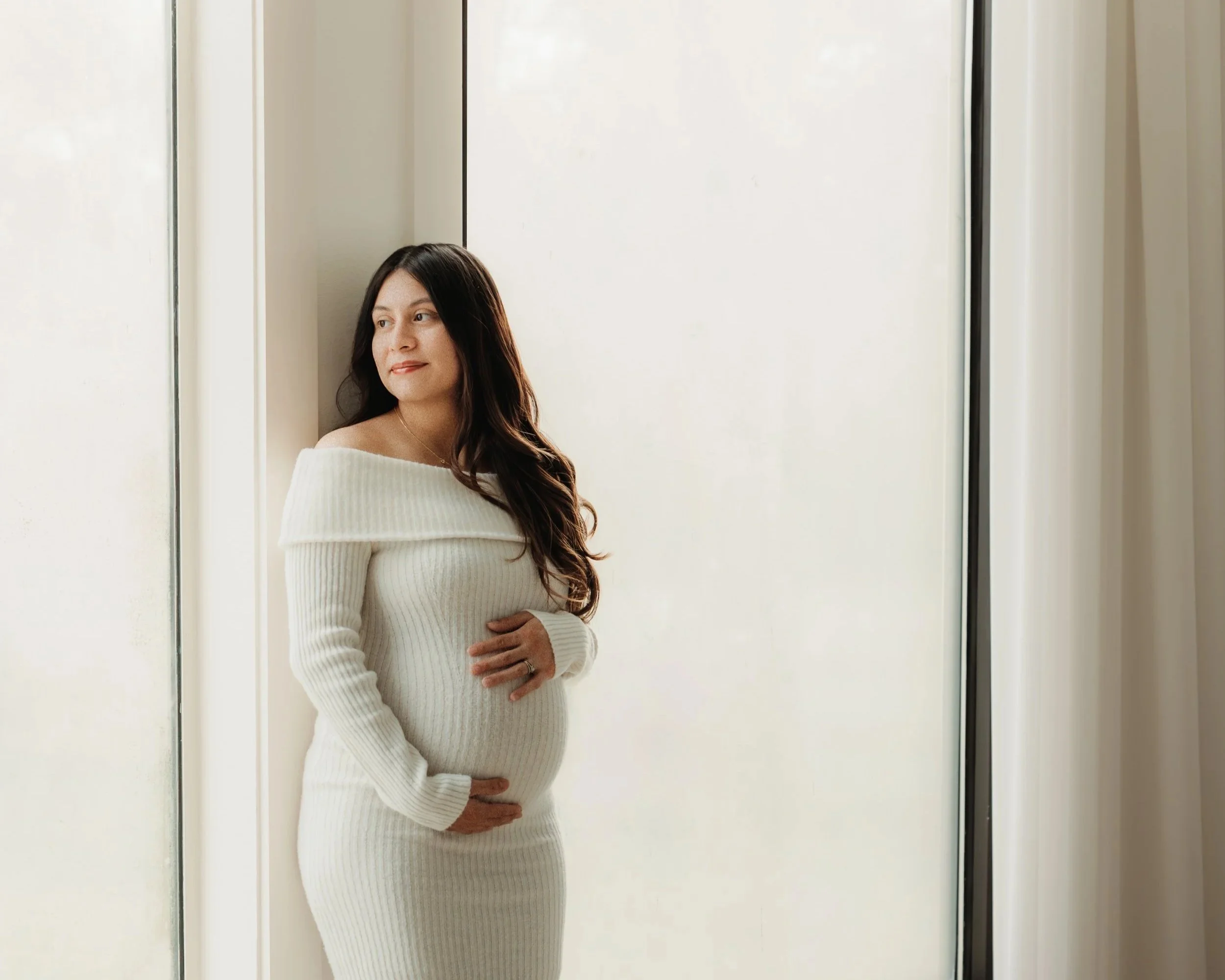 A pregnant woman with long dark hair standing near a window in a white off-shoulder sweater dress, gently touching her belly and looking out thoughtfully.