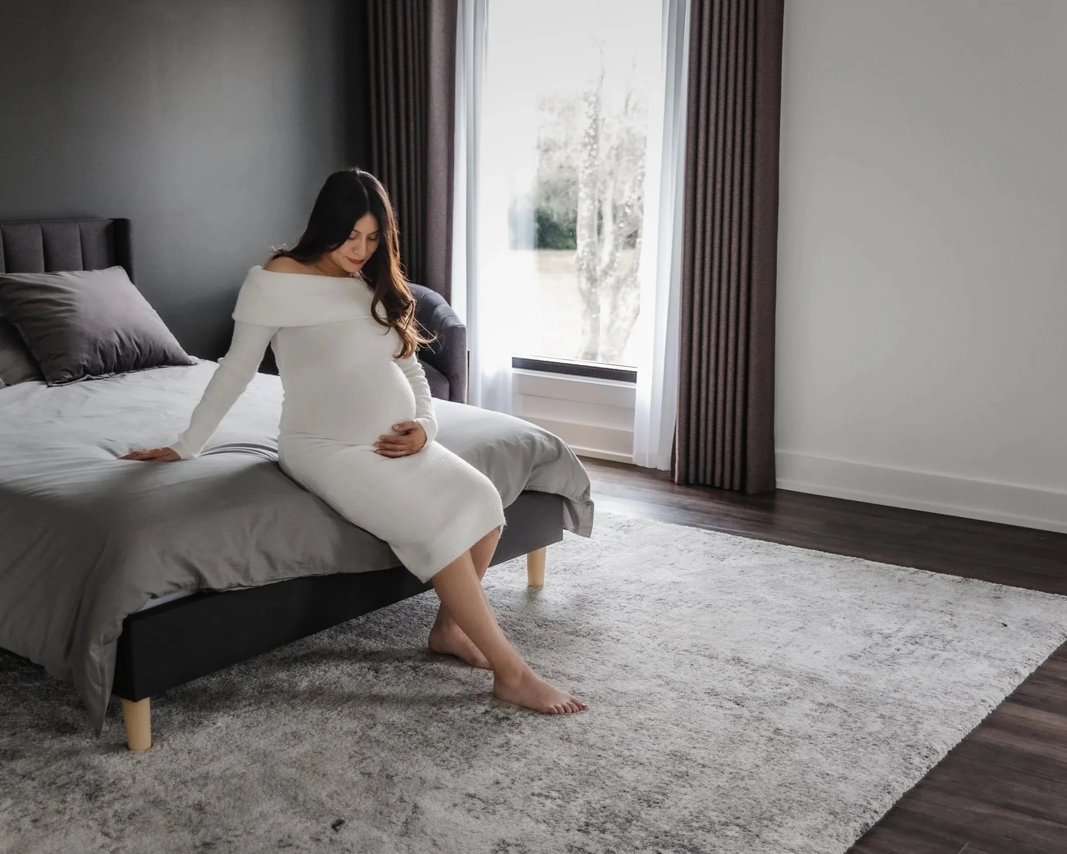 A pregnant woman in a white dress sitting on the edge of a bed in a modern bedroom, touching her belly gently with her right hand, looking down while resting her left hand on the bed, with a large window with curtains behind her and a soft-colored ru