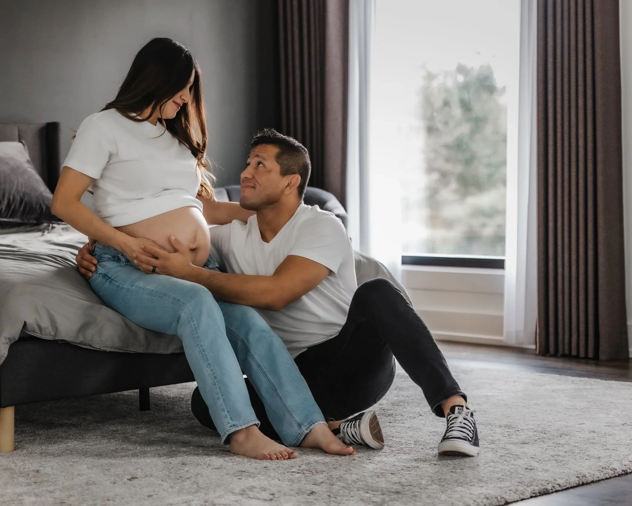 A couple with a pregnant woman sitting on a bed in a bright, modern room. The man is kneeling before her, looking up lovingly, with his hands on her pregnant belly. The woman is gently holding her belly and smiling down at him.