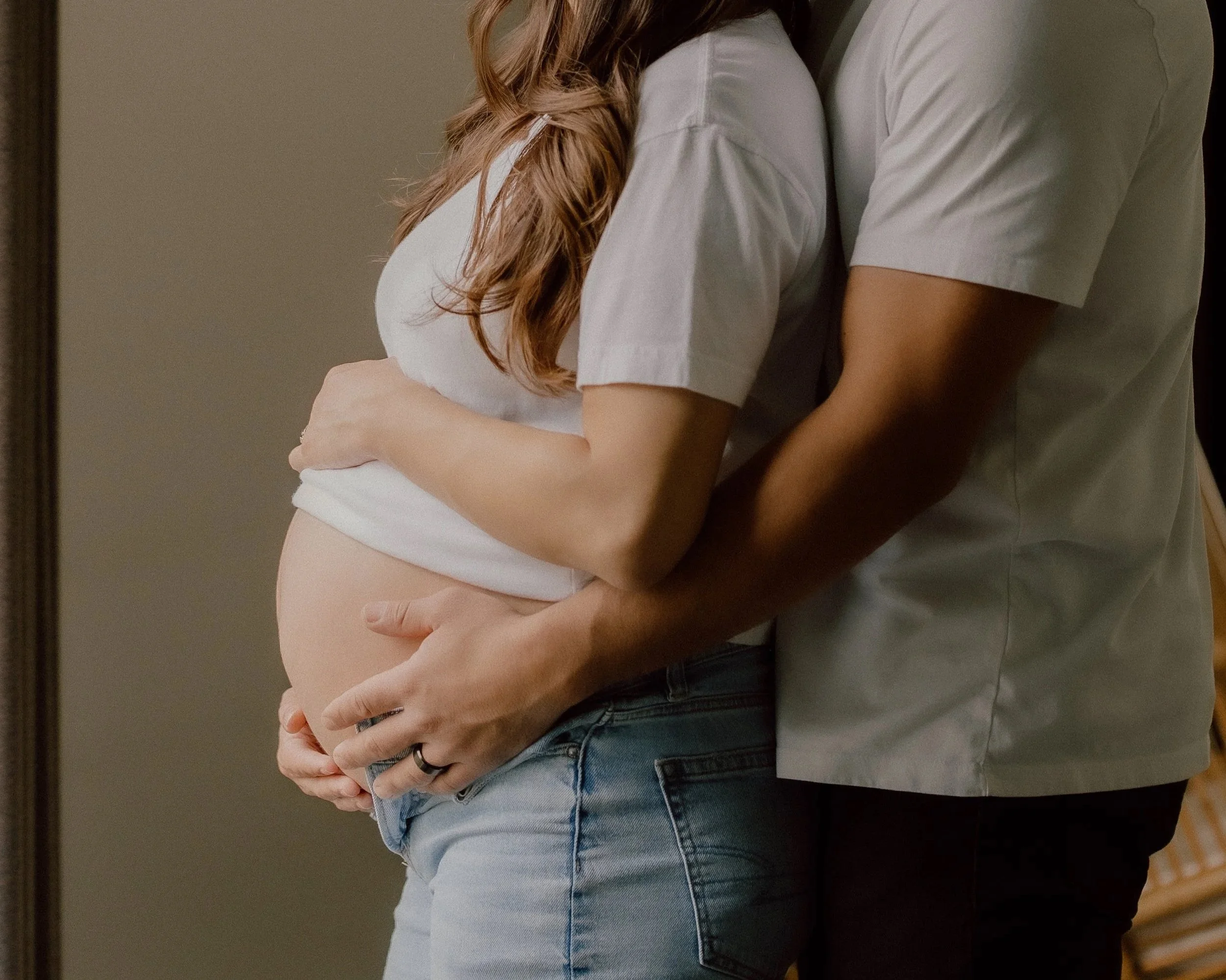 Couple embracing, with woman showing her pregnant belly, both in casual white shirts and jeans.