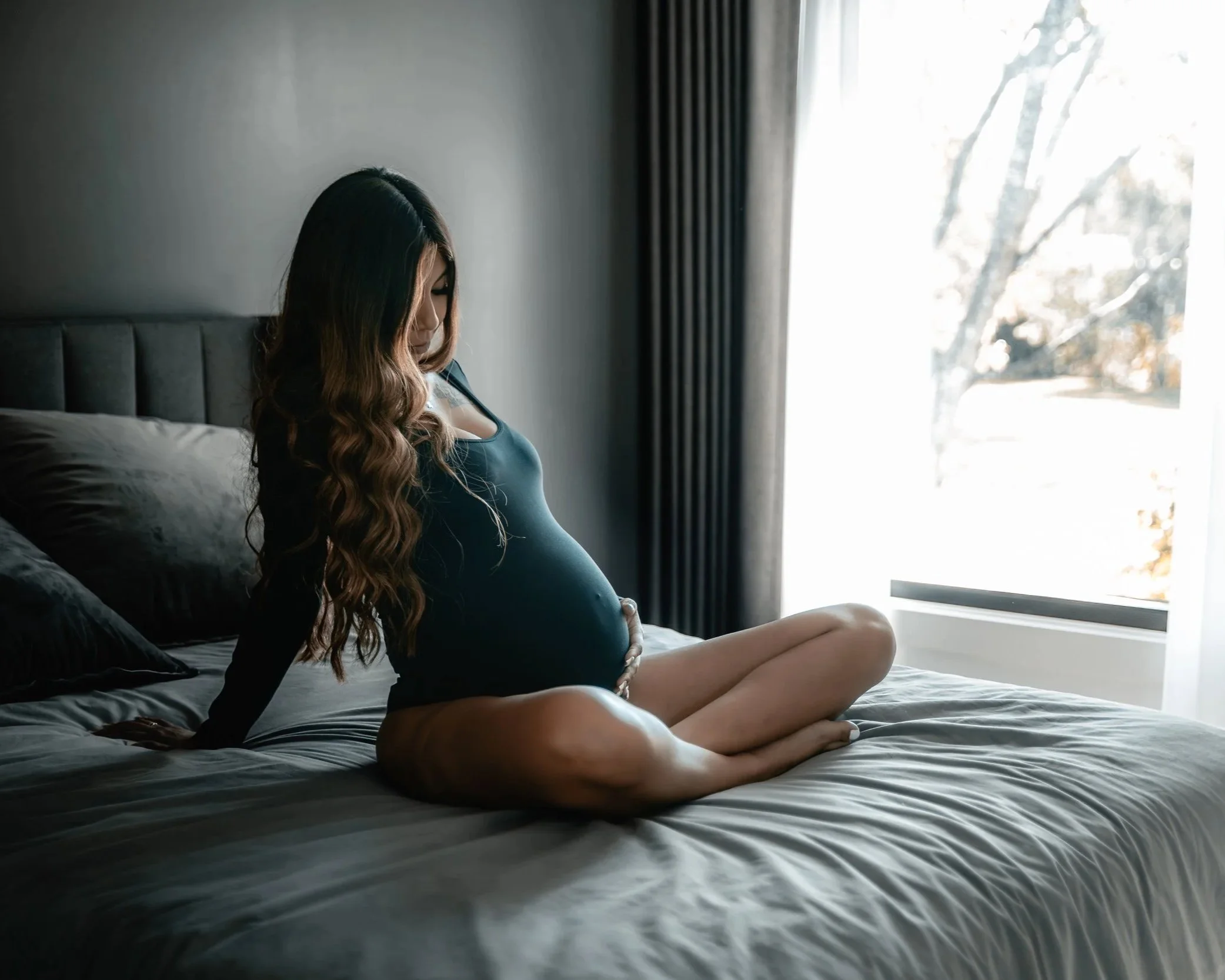 A pregnant woman sitting on a bed in a dimly lit room near a window with sunlight streaming in, looking down at her belly.