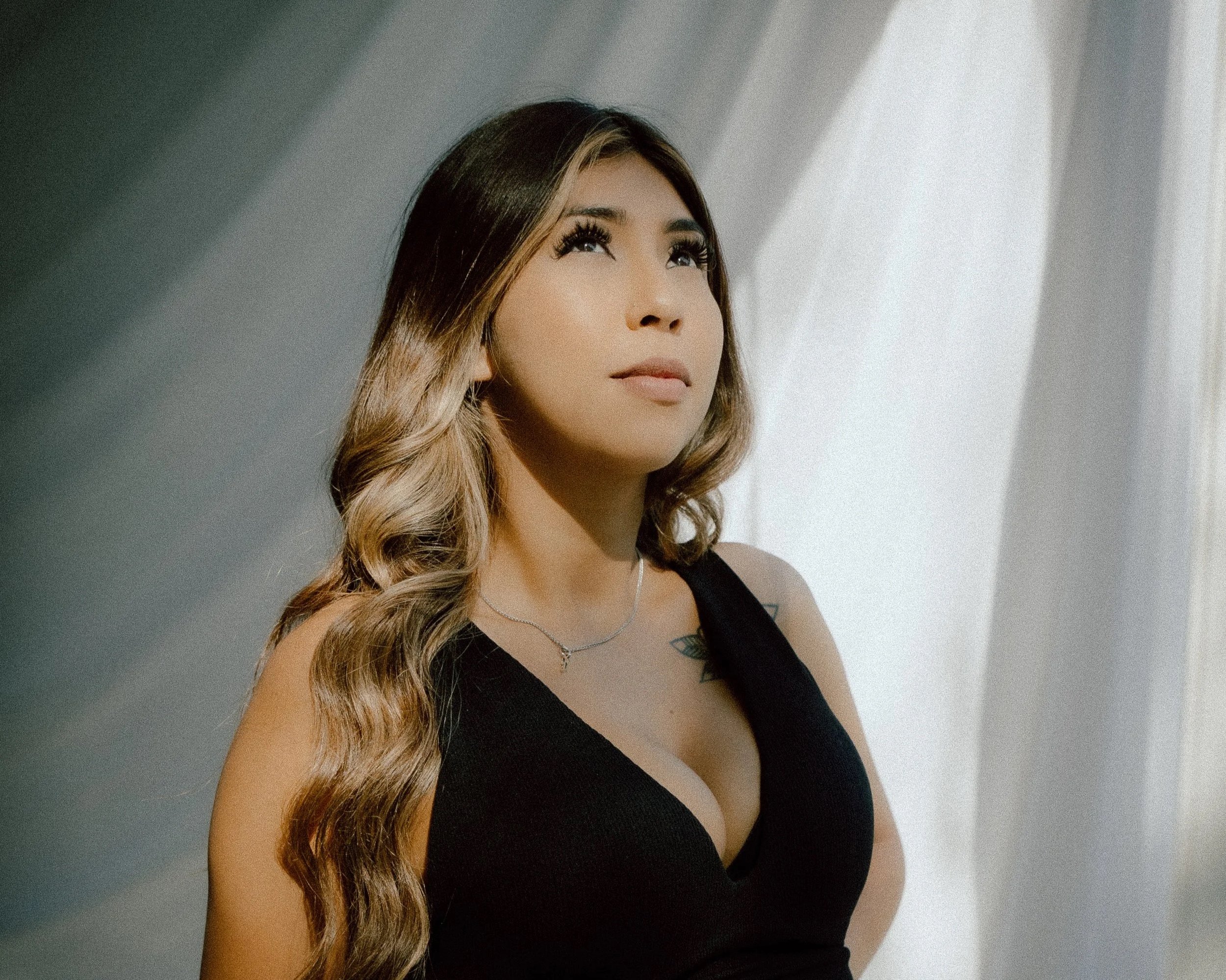 A woman with long, wavy hair looking upwards, wearing a black sleeveless top and a necklace, standing in front of a white wall with shadows.