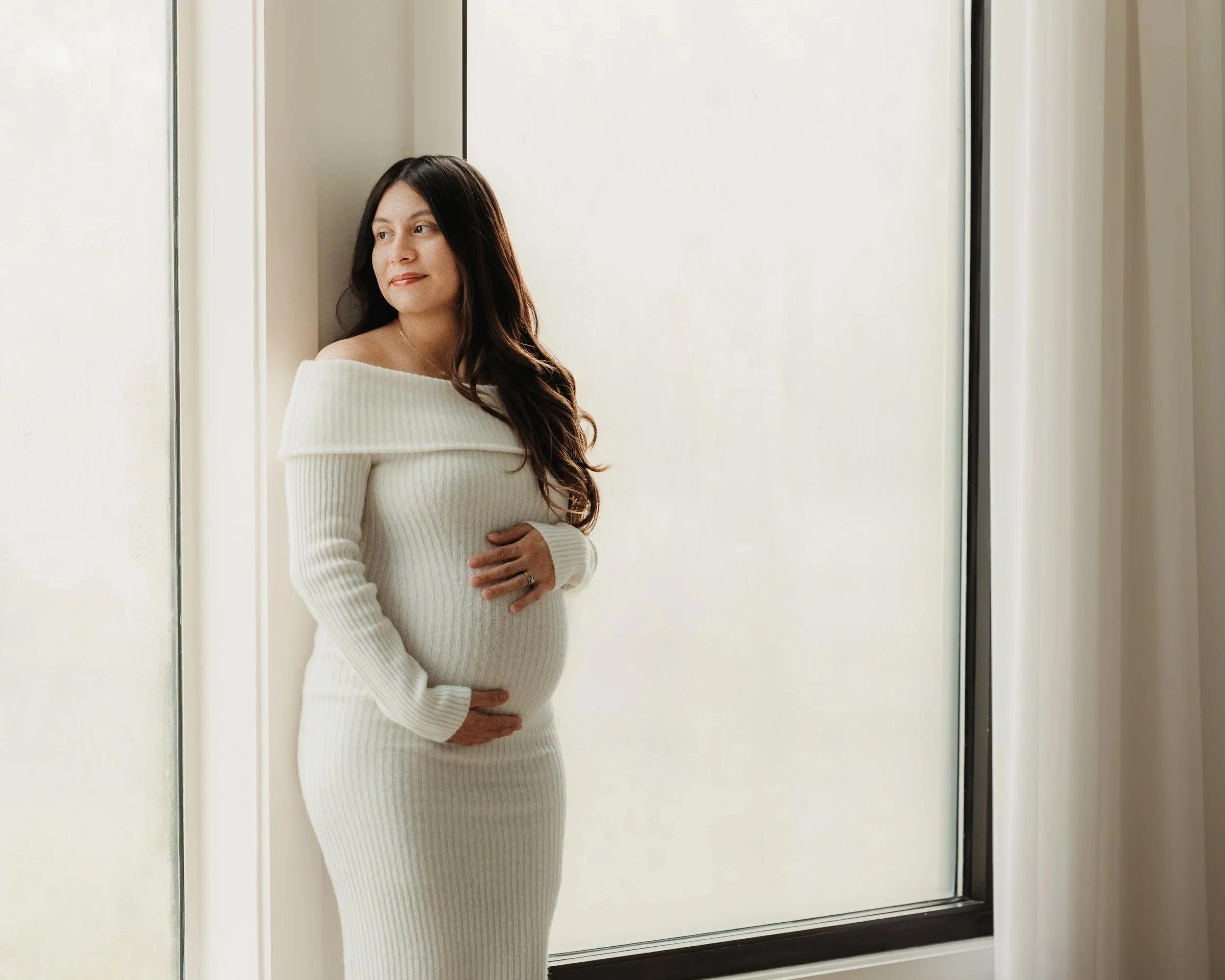 Pregnant woman in a white knit dress standing by a window, gently touching her belly, looking thoughtfully outside.
