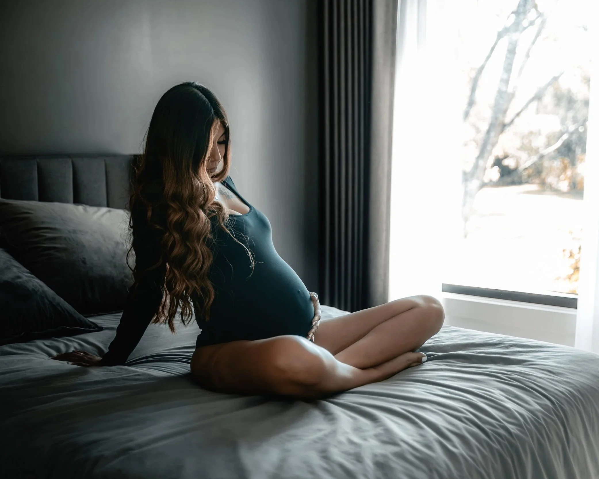 A pregnant woman sitting on a bed near a large window with curtains, gently holding her belly with her hand, looking down in a dimly lit bedroom.