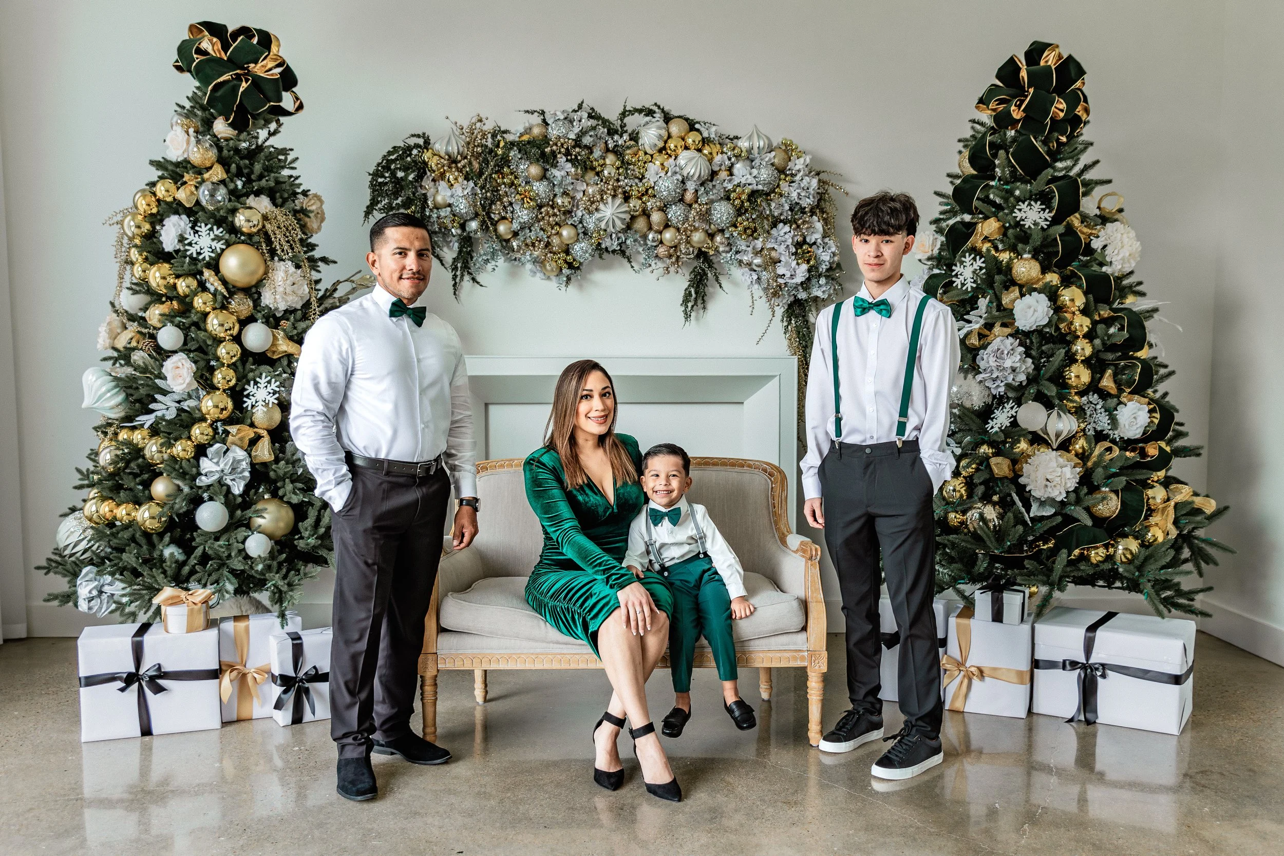 A family of five celebrating Christmas indoors, with decorated Christmas trees and wrapped presents. The family members are dressed in formal attire, with the woman and boys wearing matching green and white outfits. The woman is seated on a vintage s