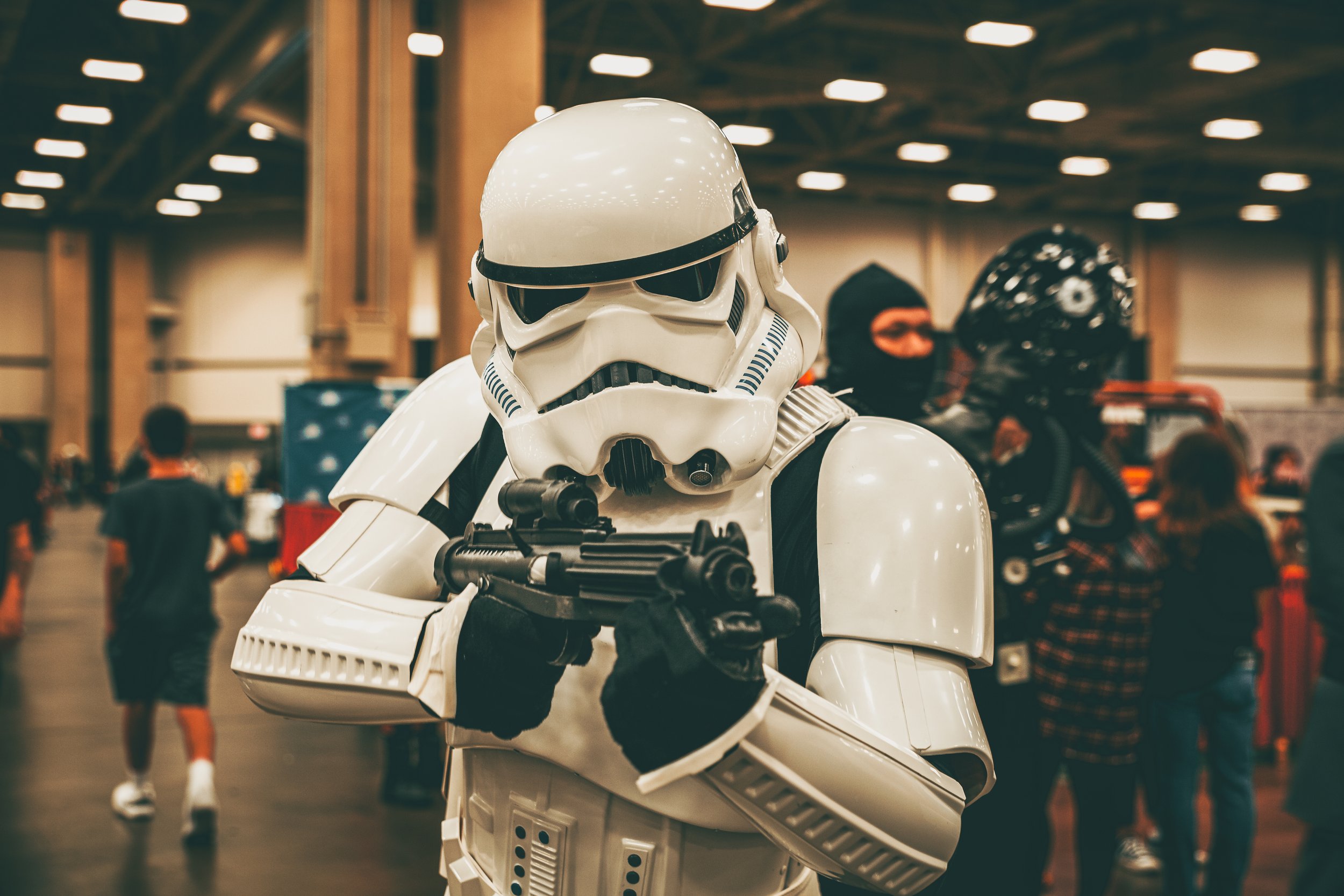 A person dressed as a Star Wars stormtrooper holding a blaster, standing in an indoor event space with people and booths in the background. Comic-con Star Wars