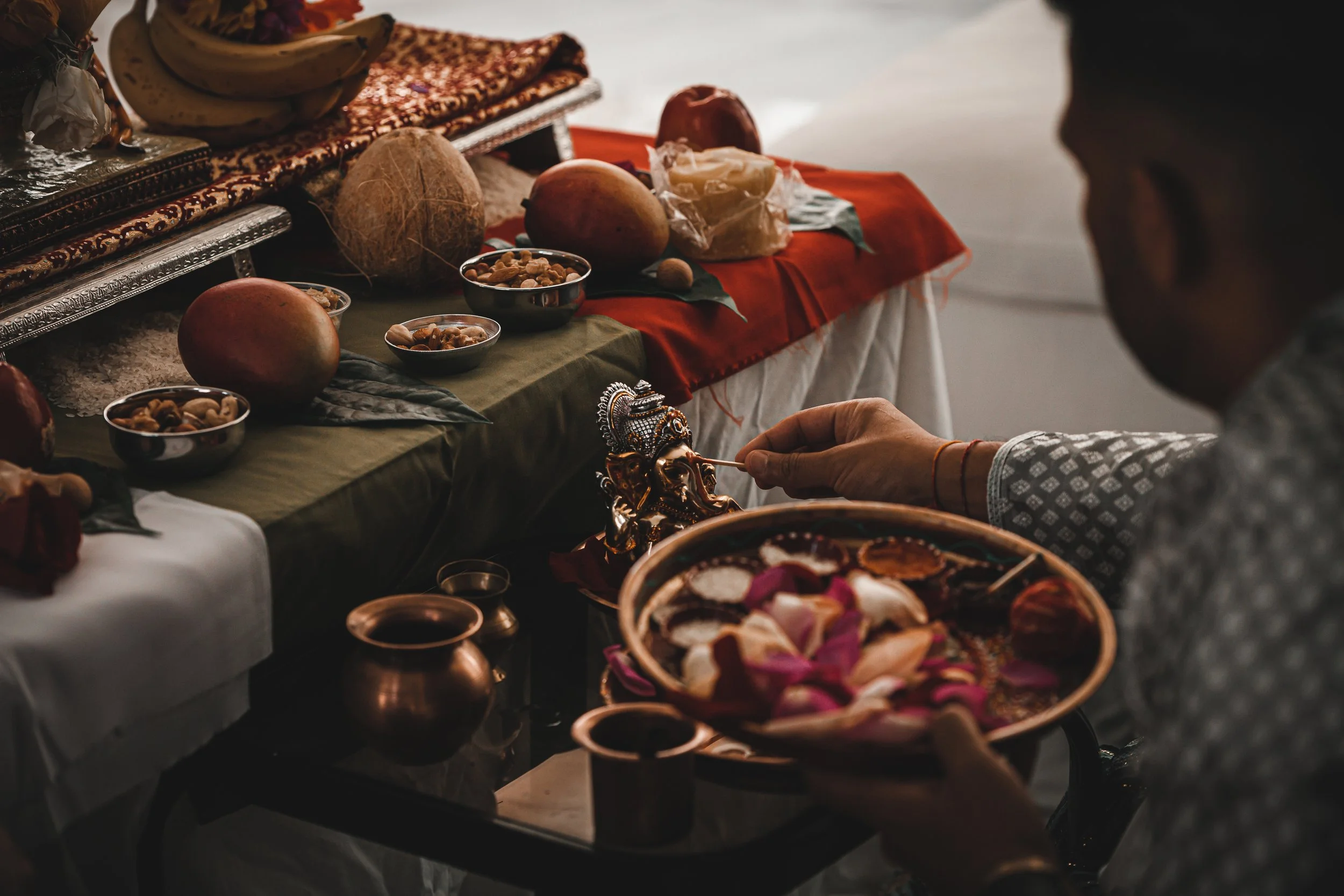 A person performing a Hindu or Hindu-related religious ritual, holding a decorated tray with flower petals, in front of a table with various fruits, nuts, and religious items like a small idol and vessels.