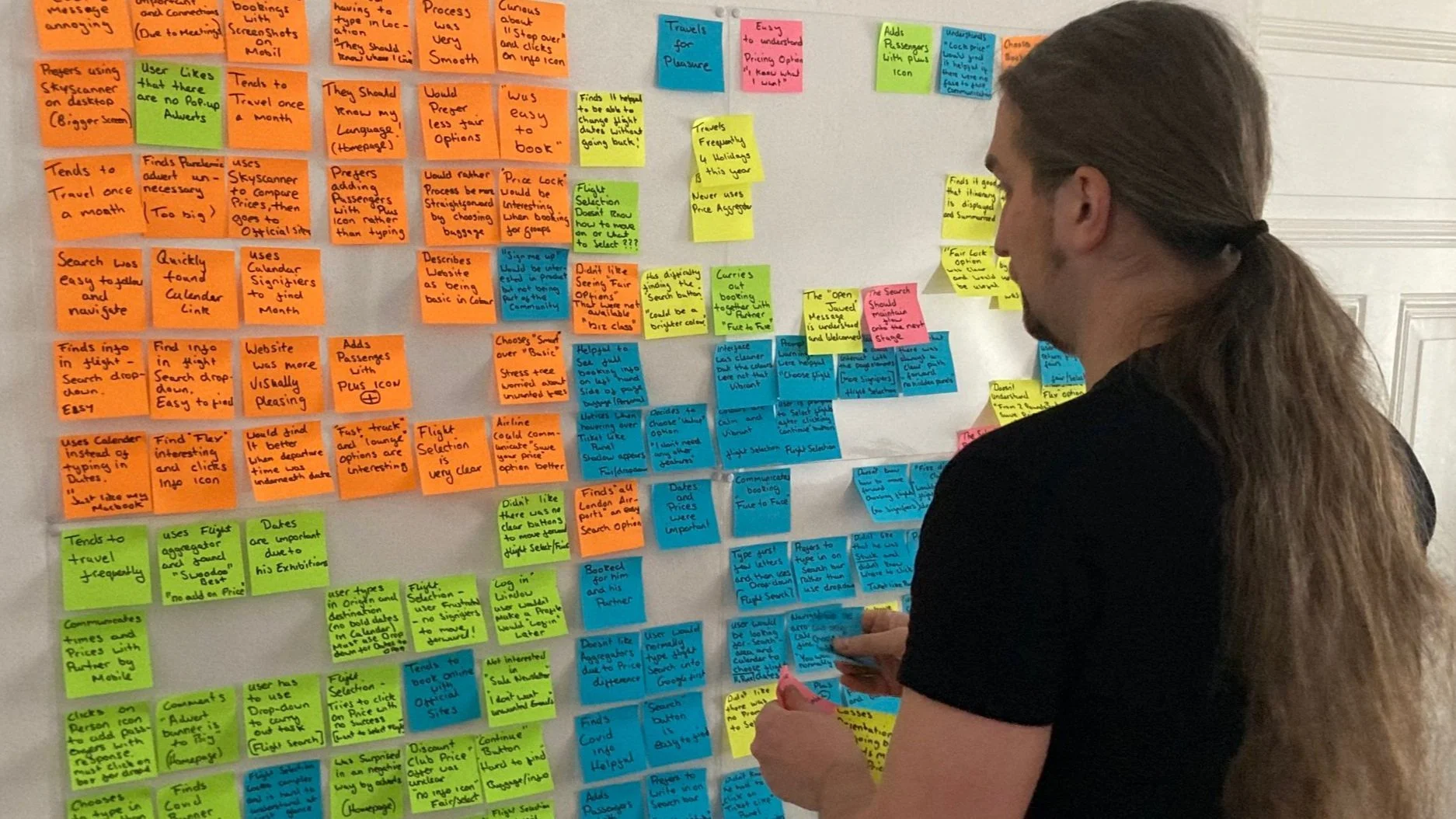 A woman with long hair tied back in a ponytail and wearing a black shirt, standing in front of a wall covered with multiple colorful sticky notes arranged in columns. She appears to be writing or organizing ideas on the notes.