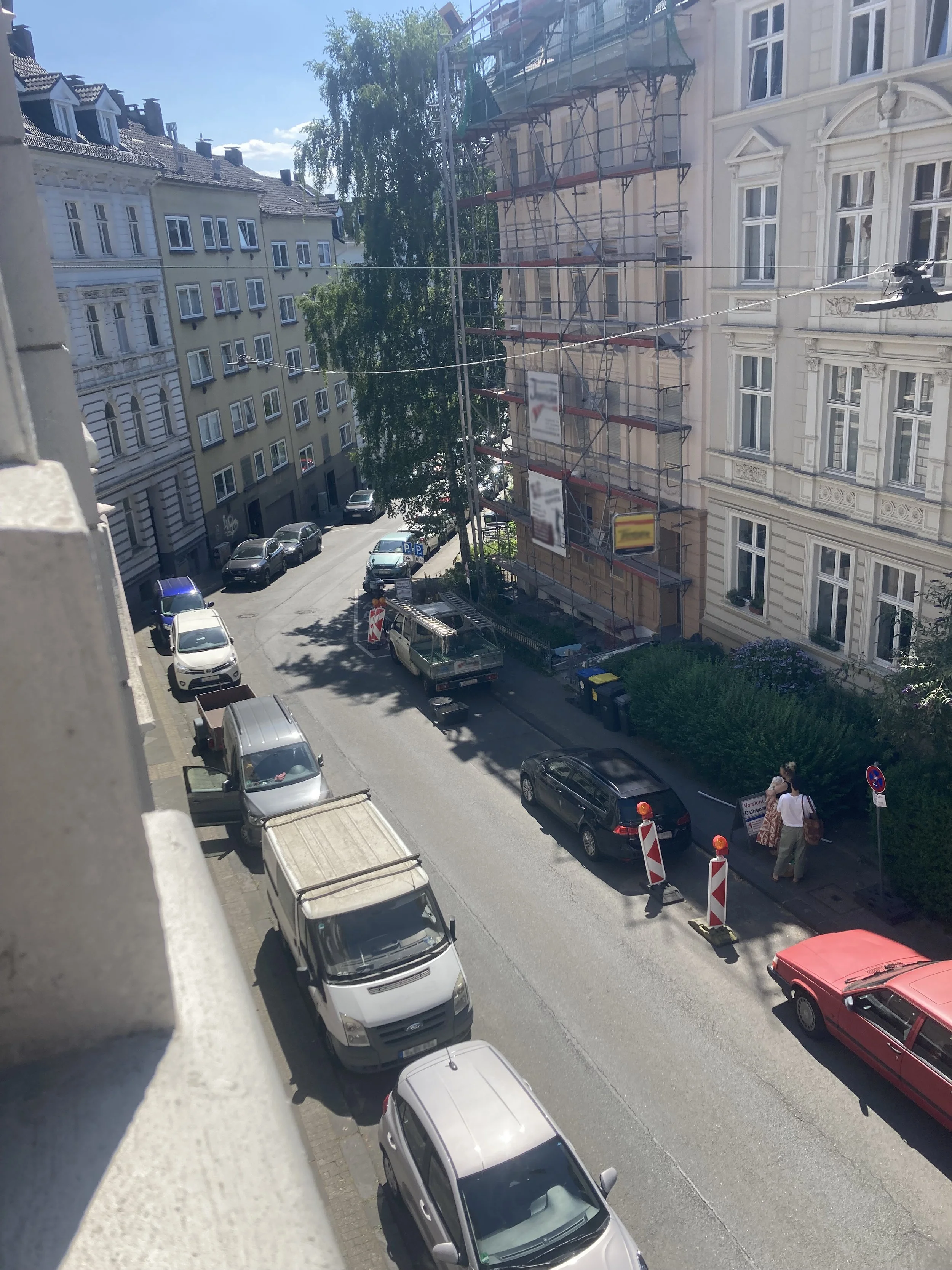 A street scene with parked cars along a building under renovation. The building is covered in scaffolding, and there are residential buildings, trees, and pedestrians visible in the background on a sunny day.