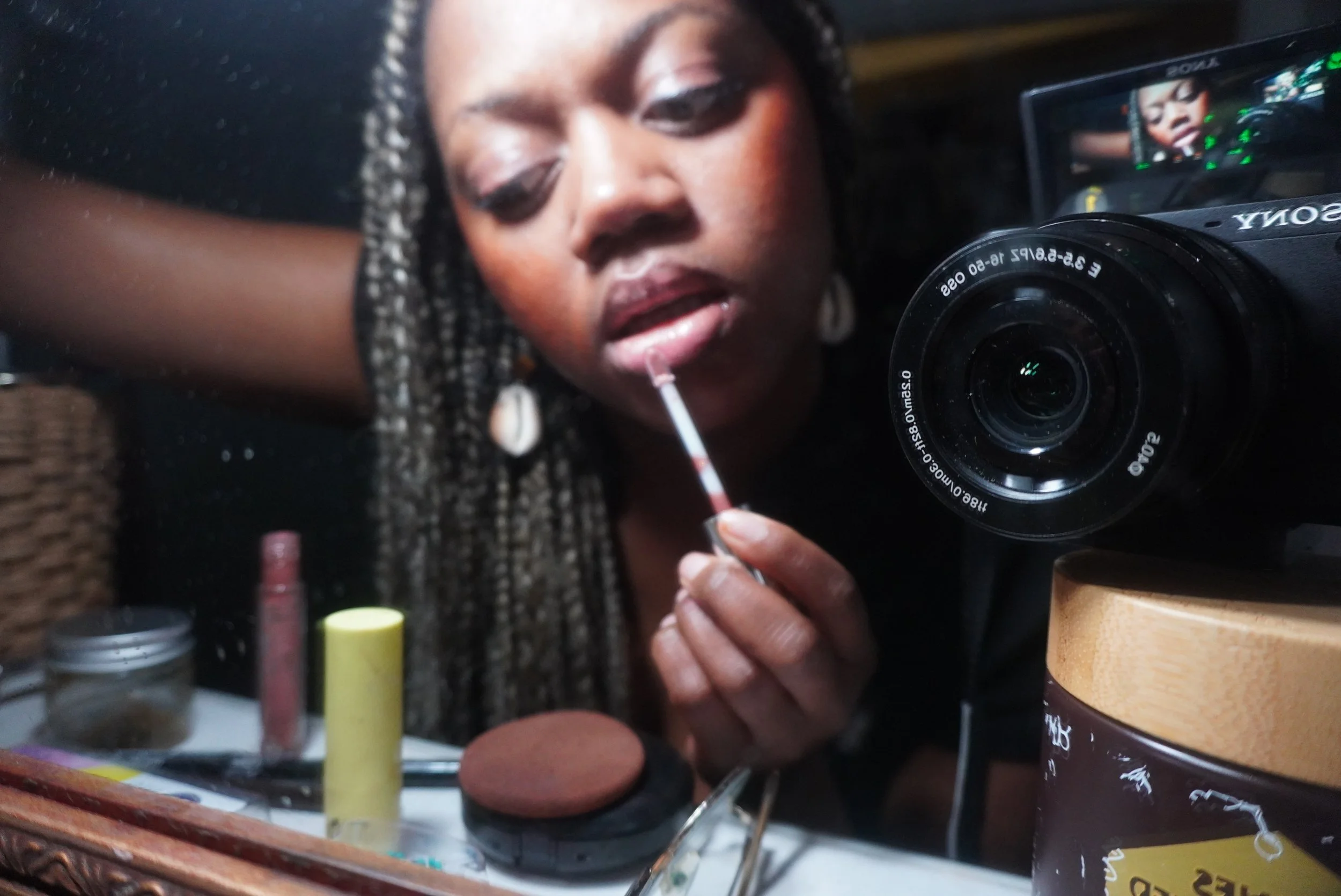 A woman applying lipstick in front of a mirror with a camera taking her picture.