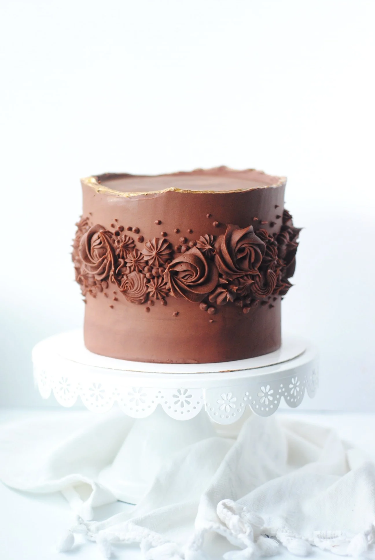 A white frosted birthday cake decorated with colorful sprinkles on top and around the sides, placed on a glass cake stand with a ruffled white cloth underneath.