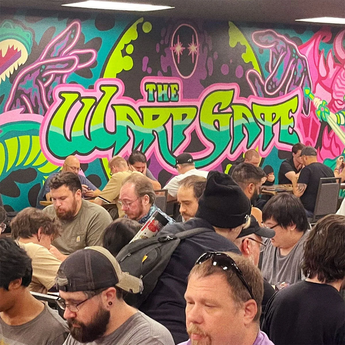 The Warp Gate logo mural in green and pink above players at tables in the Columbus, Ohio card shop.