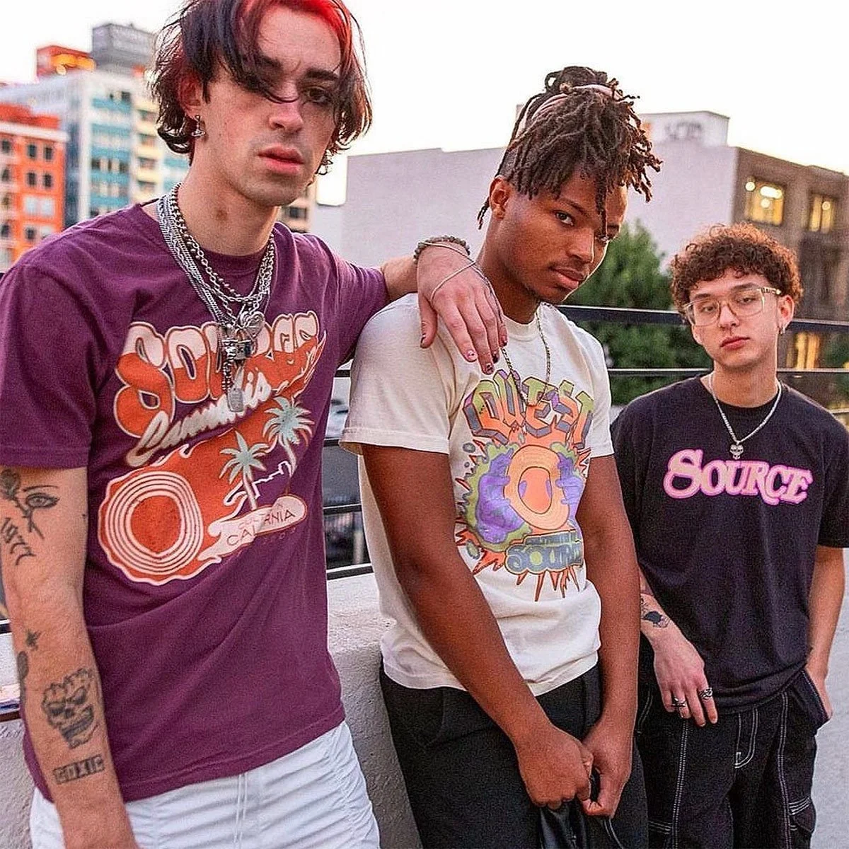 Three people wearing Source Cannabis graphic tees in maroon, cream, and black on a rooftop at dusk.