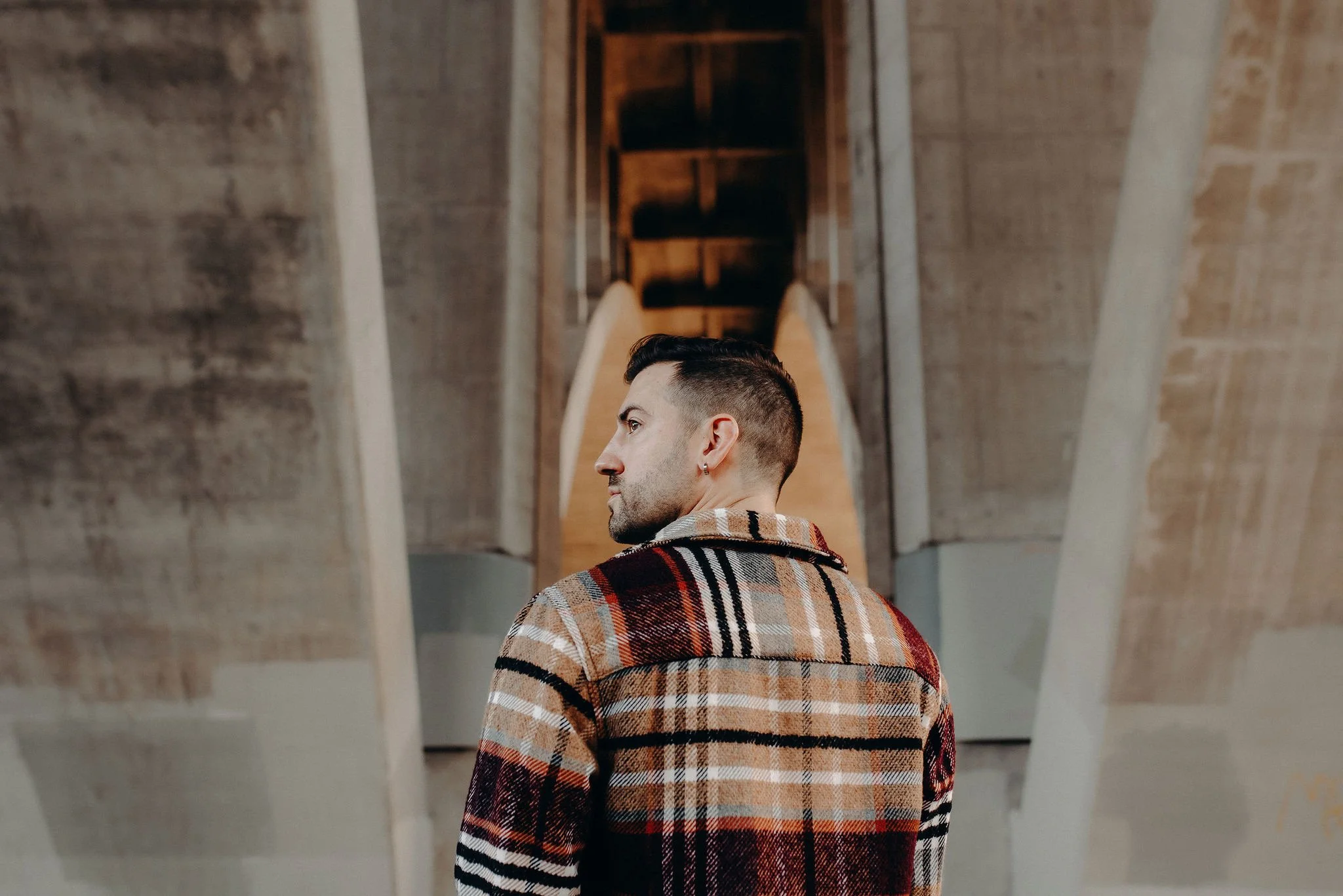 A man with a short hairstyle and a small earring wearing a plaid shirt, looking over his shoulder in an industrial-style interior with wooden and concrete architectural features.