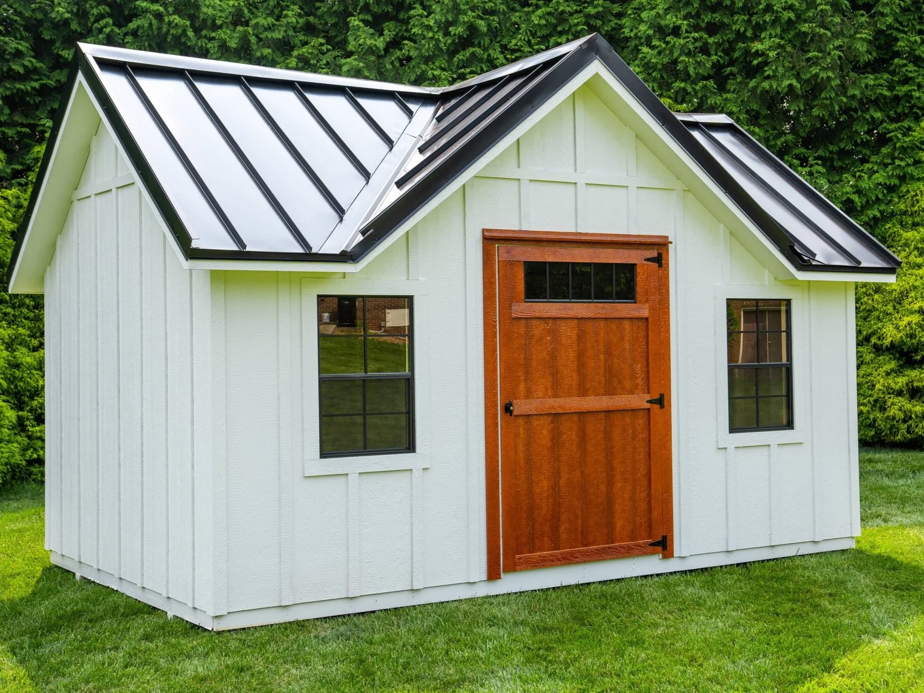 A small white shed with black roof, wooden door, and windows, situated on a grassy lawn with trees in background.