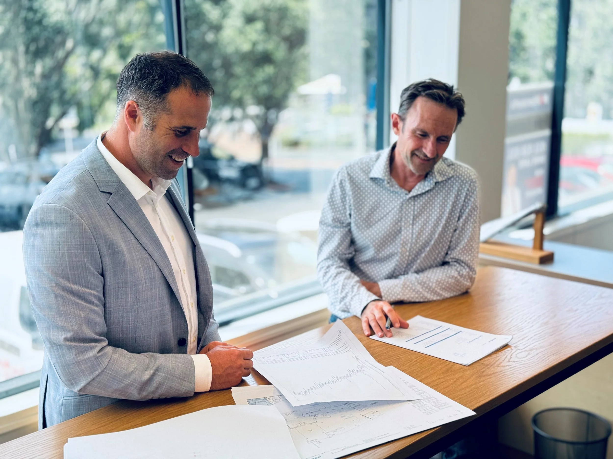 Two men in business attire smiling and reviewing documents on a wooden table inside a well-lit office with large windows.