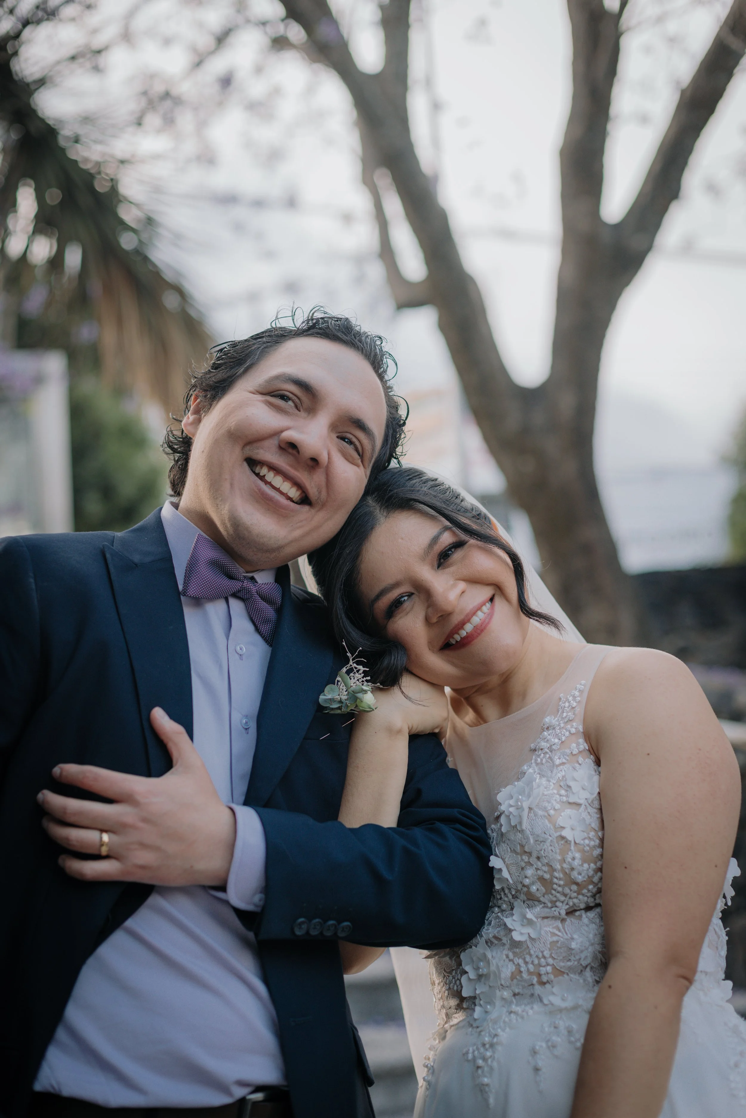 Pareja en vestido de boda sonriendo y abrazados, en un entorno al aire libre con árboles, el hombre lleva un saco azul oscuro y corbata morada, la mujer lleva un vestido blanco con encaje y flores.