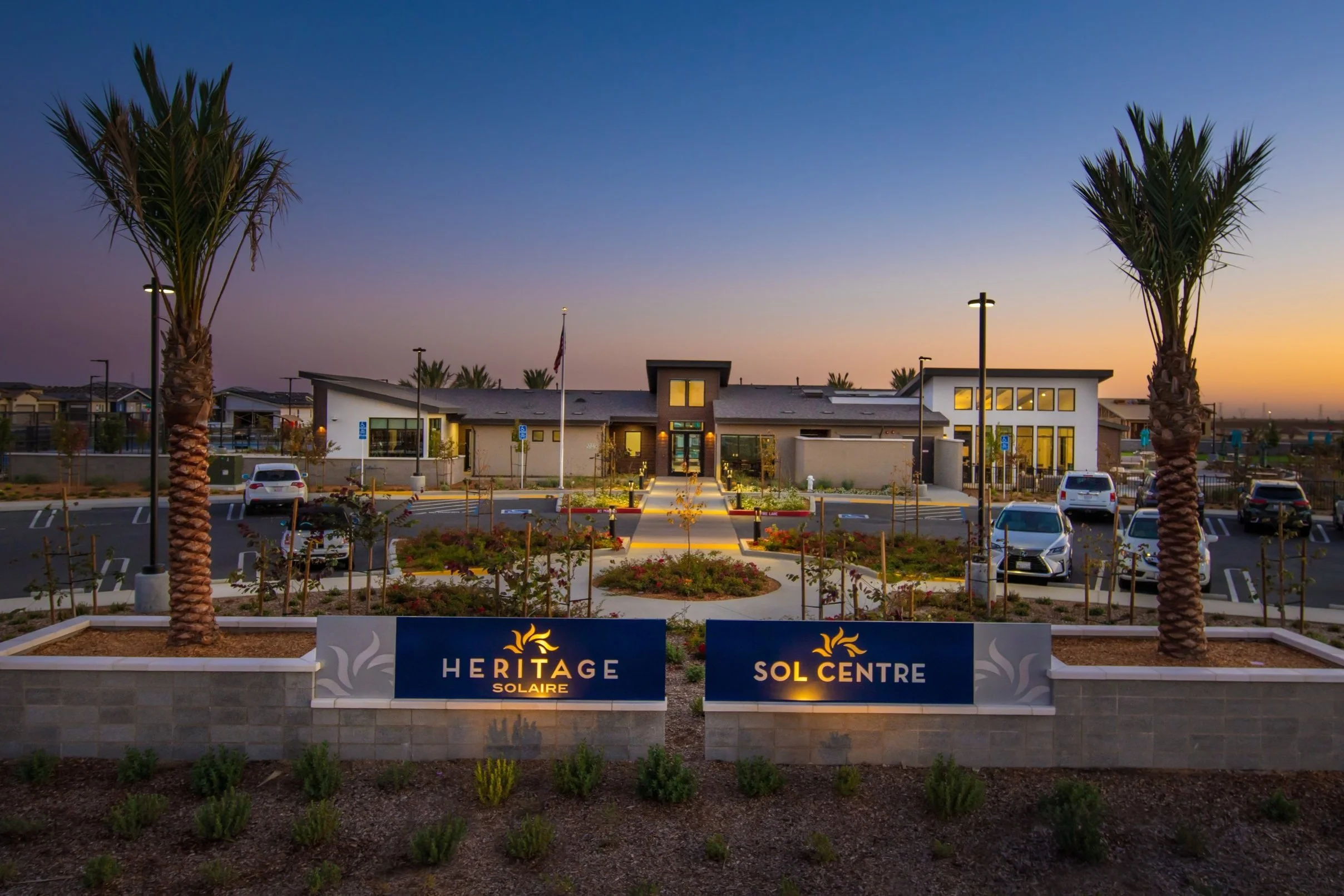 Exterior view of Heritage Solaire Sol Centre at dusk with a landscaped entrance, palm trees, and a parking lot in front.