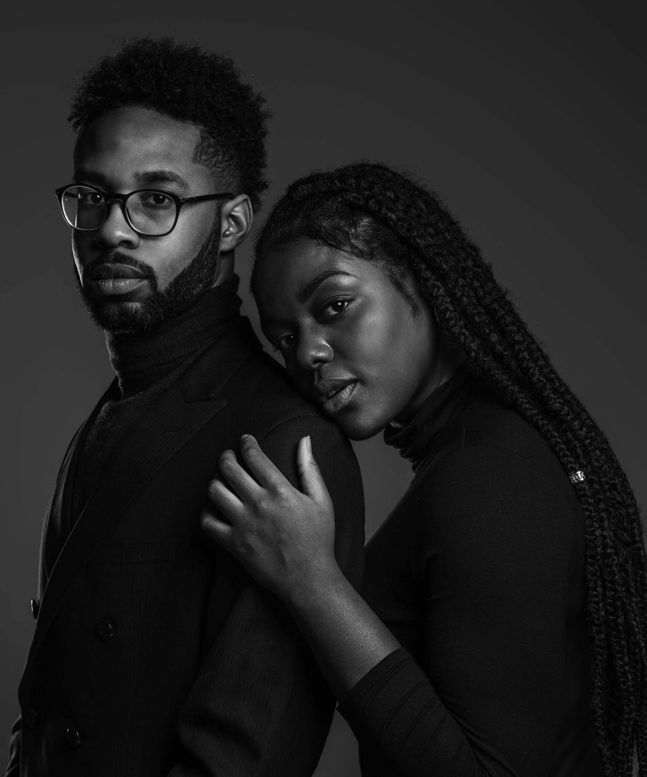 Black and white portrait of a man with glasses and a woman with braided hair in a close pose, both wearing dark clothing.