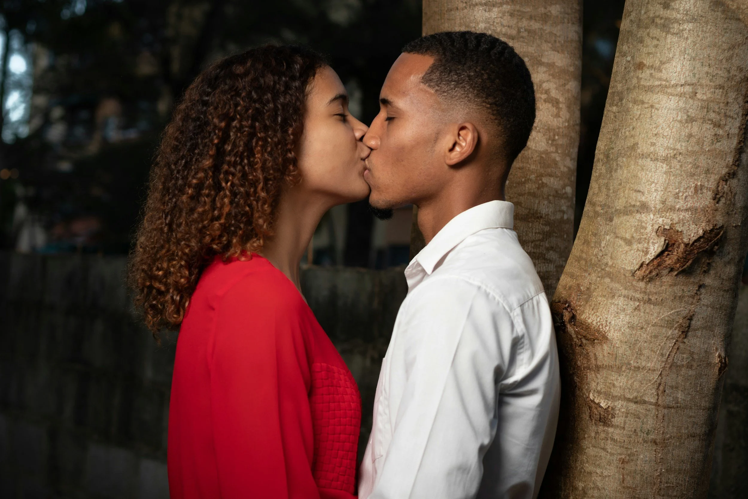 A couple kissing near trees, woman in red and man in white shirt