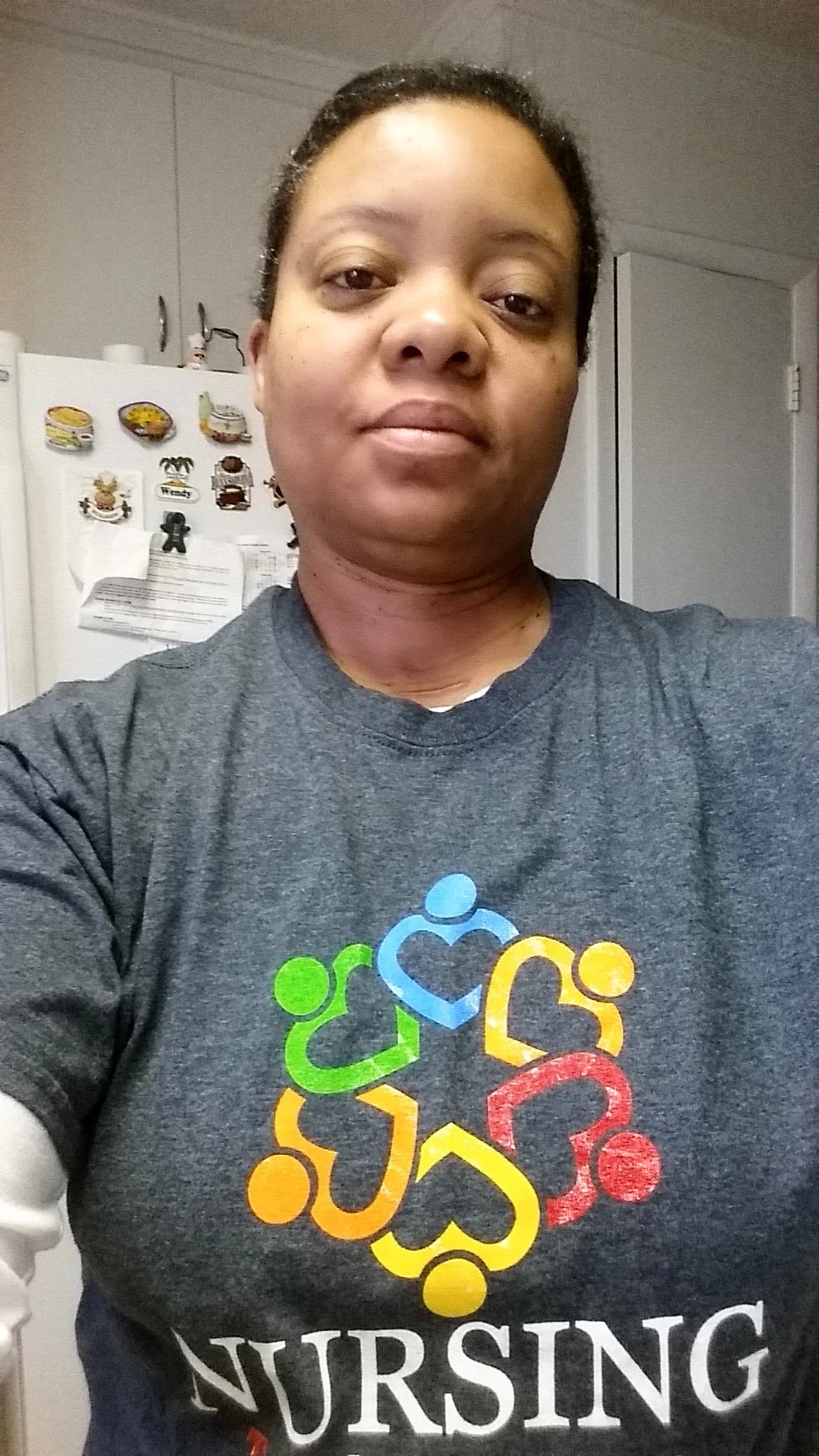 A woman taking a selfie in a kitchen, wearing a dark gray shirt with colorful nursing-themed logo and the word 'NURSING' on it.
