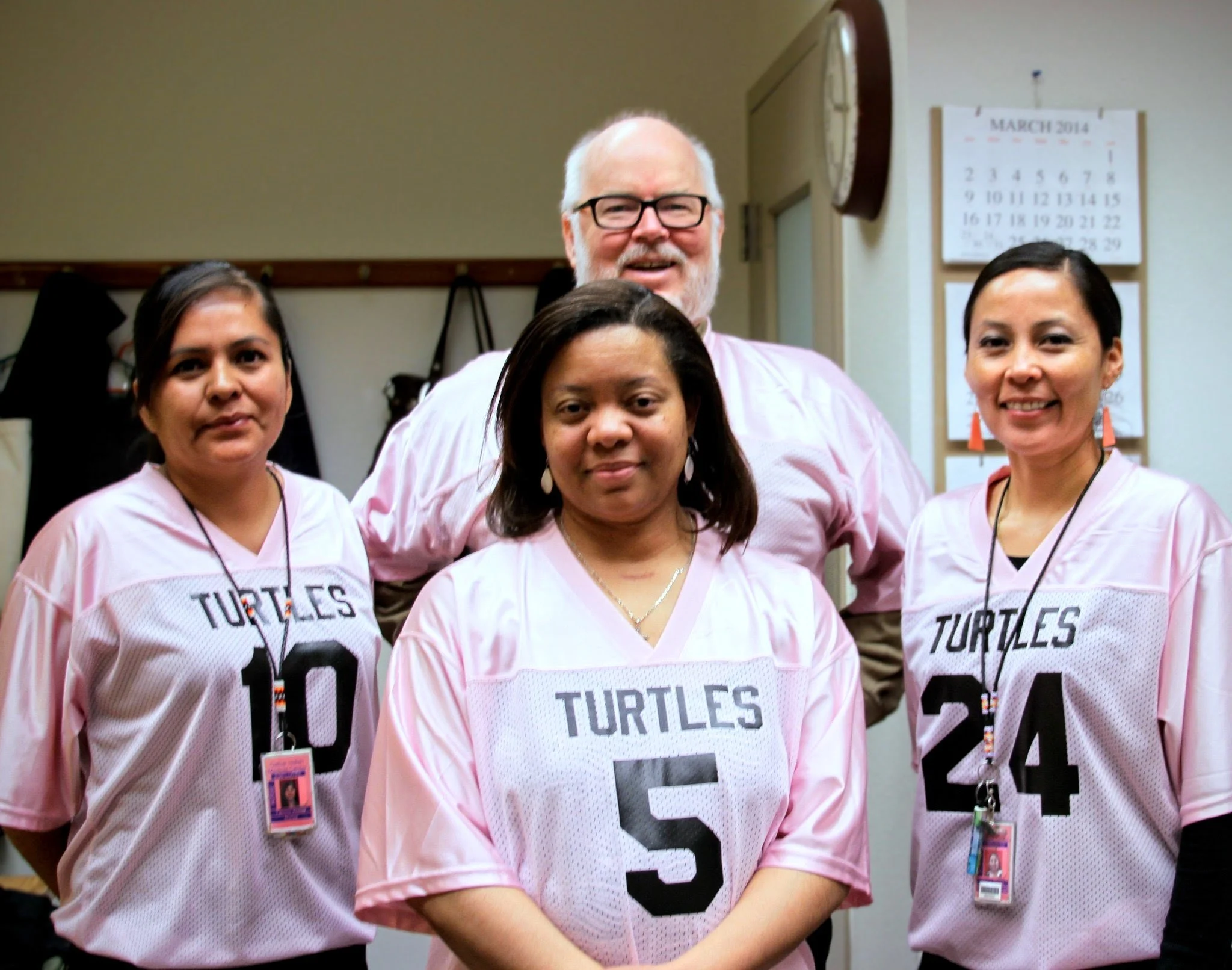 Group of four diverse women wearing pink sports jerseys with the word "TURTLES" and numbers, smiling in a room with a wall clock and calendar.
