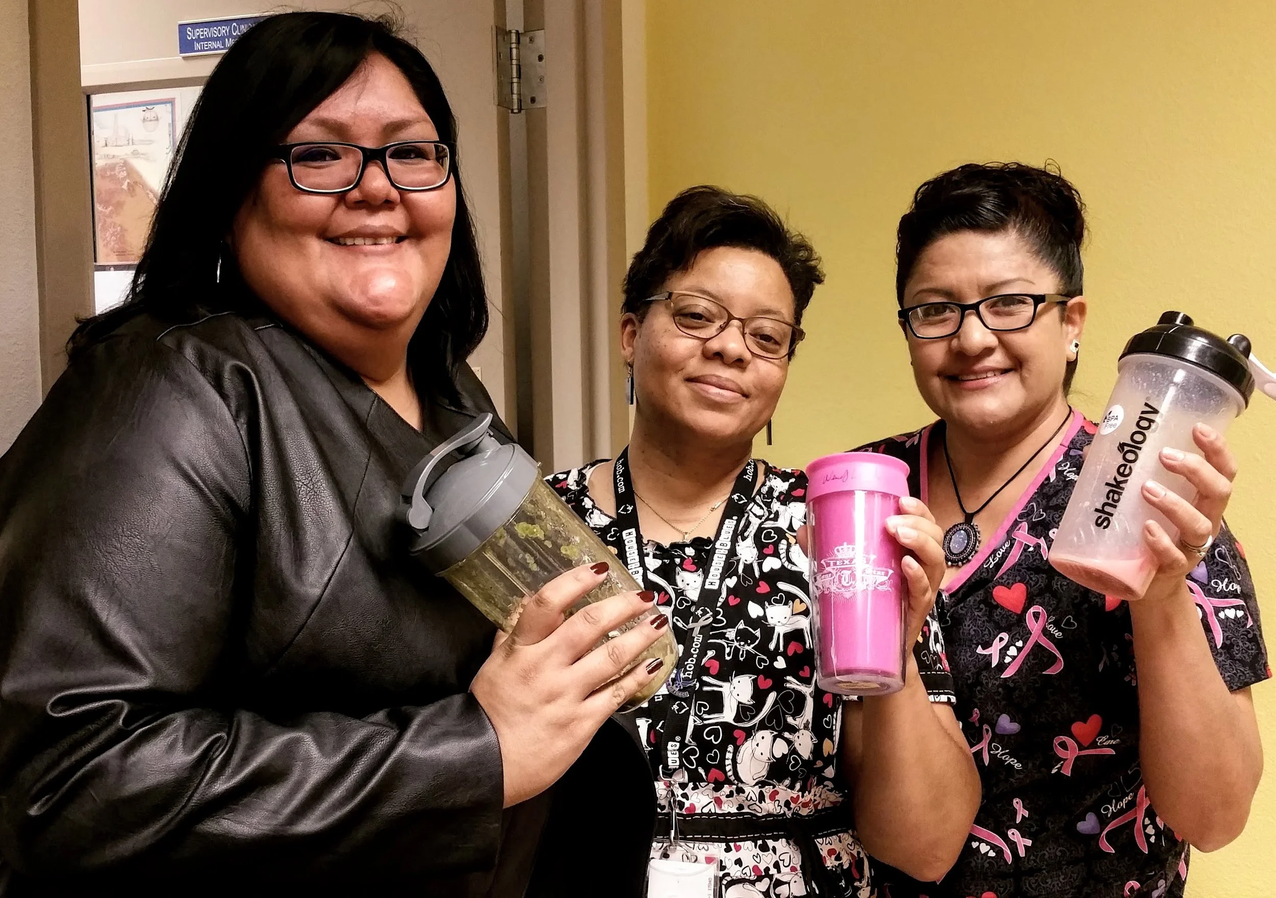 Three women standing together indoors, holding reusable drink containers, wearing breast cancer awareness ribbons, smiling, and wearing patterned clothing and glasses.