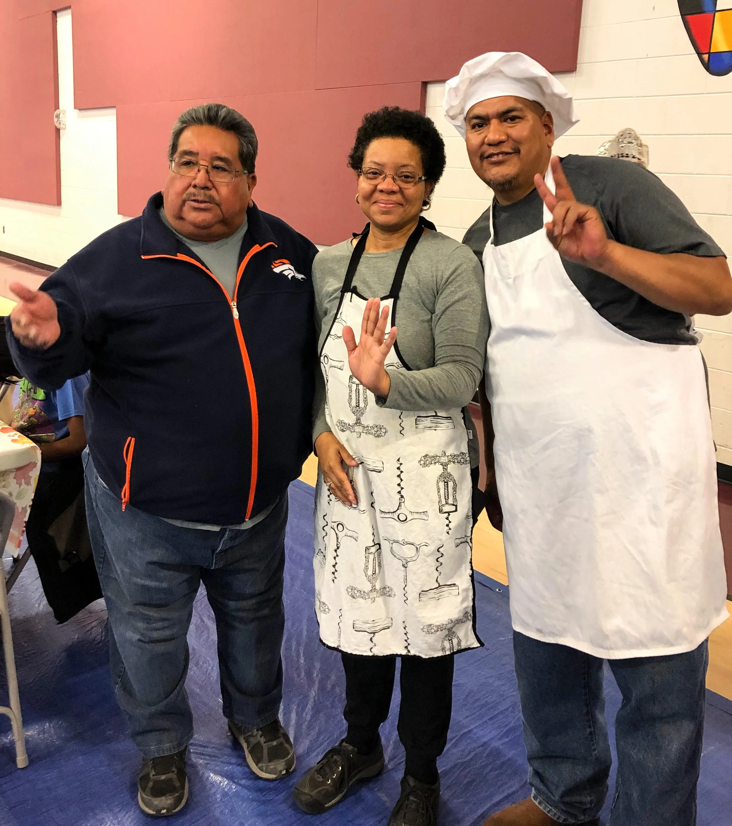 Three people posing together indoors. The person on the left is a man with glasses wearing a dark jacket with orange accents. The middle person is a woman with glasses, short curly hair, and a gray shirt with a black lanyard. The person on the right 