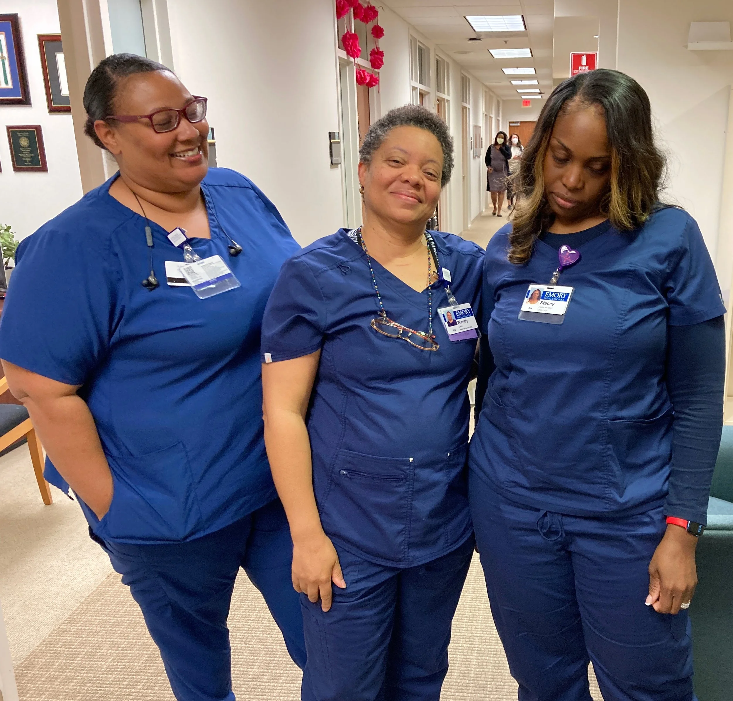 Three women in medical scrubs smiling and looking down in a hospital hallway with pink decorations and other people in the background.